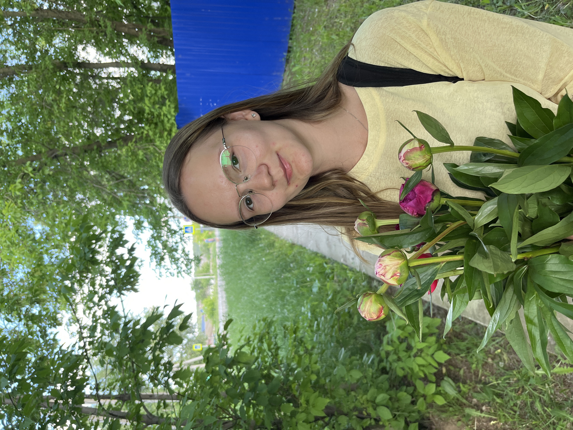 A young female, appearing to be in her late teens or early twenties, is depicted outdoors in the city of Chaikovsky, Russia. She has long, straight, light brown hair with subtle lighter streaks, styled with a middle part. She wears round, silver-rimmed glasses and a small, clear-stoned stud earring in her right ear. A thin silver chain necklace is visible on her neck. Her attire consists of a light yellow, long-sleeved, crew-neck top, with a black strap from a bag or backpack visible over her right shoulder. She holds a bouquet of fresh peonies, primarily consisting of closed green buds with red/pink tips, along with one partially opened magenta peony bloom and several large green leaves. Her facial expression shows a slight, closed-mouth smile directed towards the viewer.
The background environment is characterized by abundant greenery. To the subject's left, a concrete paved path runs parallel to a dense array of green bushes and trees. Beyond this immediate foliage, a wider expanse of green grass or low vegetation is visible, leading to what appears to be a paved road in the middle distance. Two yellow diamond-shaped pedestrian crossing signs, each with a blue pedestrian symbol, are visible along the distant road, consistent with Russian road signage. On the far right side of the image, behind the subject, a section of a bright blue corrugated metal fence stands vertically, partially obscured by foreground foliage. The upper background features numerous green-leafed deciduous trees under a light blue sky with scattered white clouds, indicating daytime conditions.