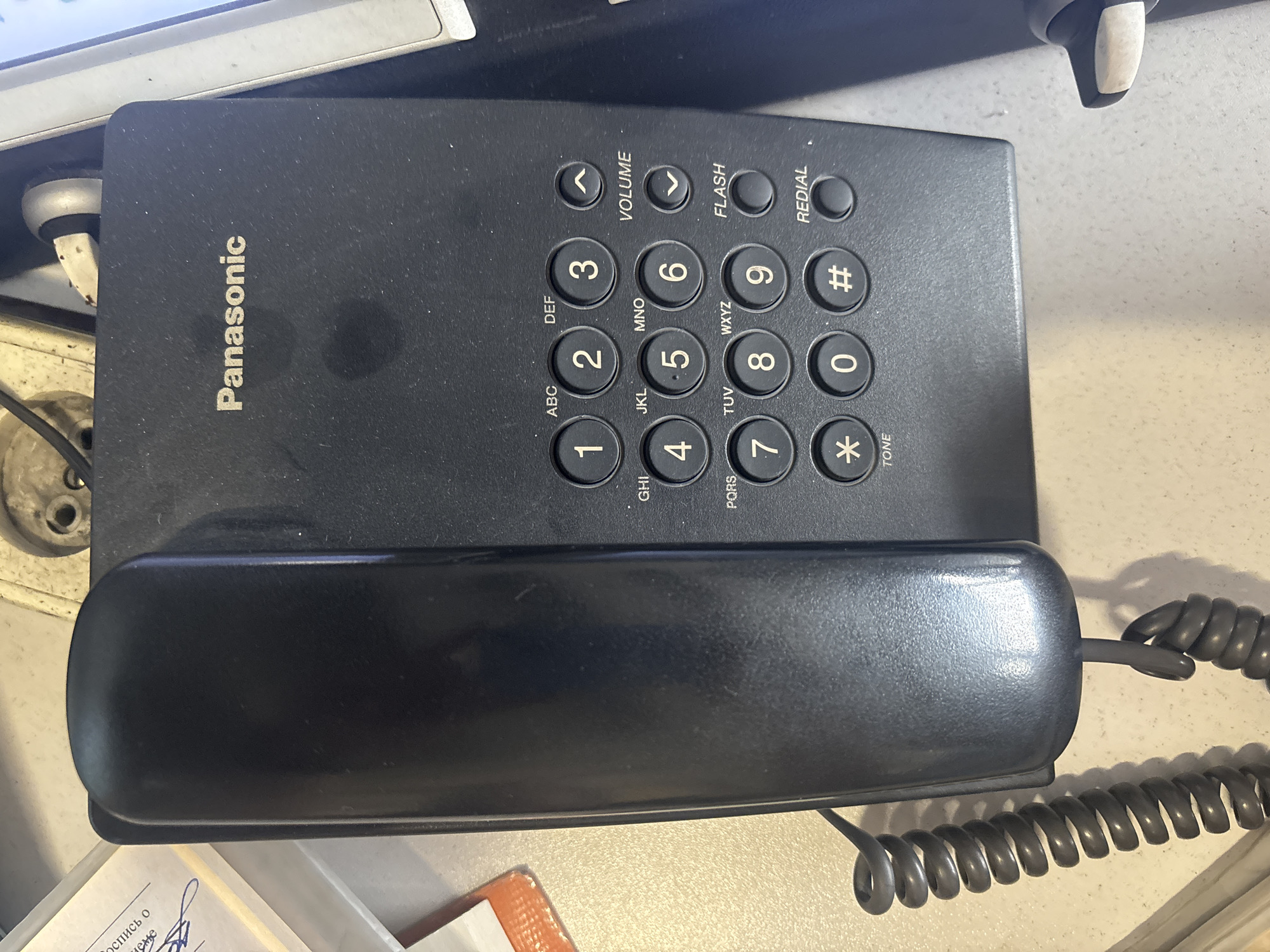 The image shows a black Panasonic corded telephone on a light-colored, textured surface, likely a desk or countertop. The telephone is oriented vertically in the frame, with the handset resting on the base. The keypad is visible, featuring numeric buttons (0-9), asterisk (*), and pound (#), along with function buttons for Volume, Flash, and Redial. The word "Panasonic" is printed in silver lettering on the upper right portion of the phone's body. To the left of the phone, a coiled cord is partially visible. A document with handwriting and a printed Russian phrase is also present in the upper left corner. The background is blurred but appears to be an indoor setting, with some electronic equipment and wires visible. The lighting suggests an overhead light source. The image is a still photograph, taken from a slightly elevated perspective. The scene is devoid of any people or discernible actions.