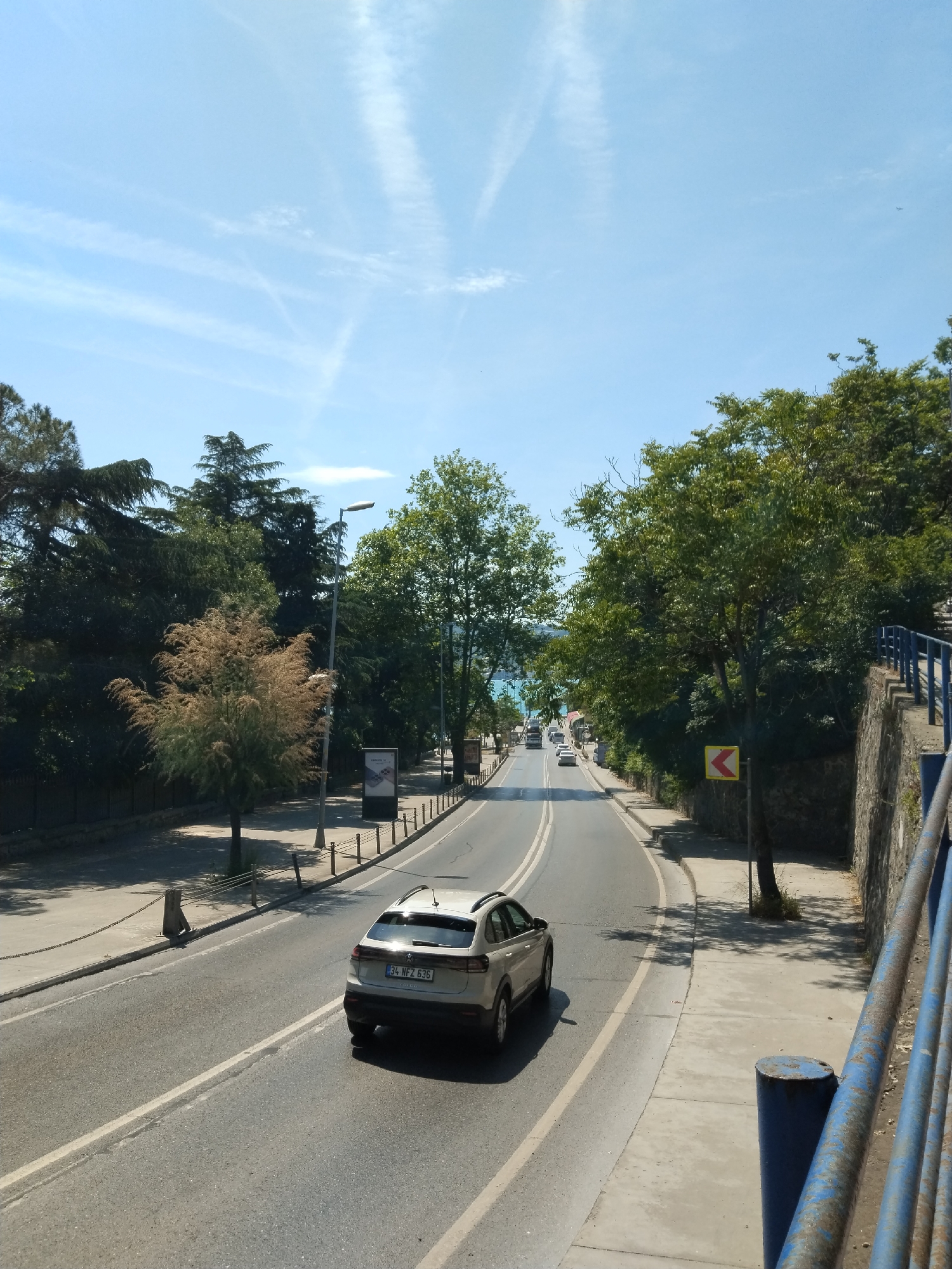 The image depicts a sunlit road scene in Sarıyer, Turkey, looking down a slightly curving asphalt road that descends towards distant water. A silver-colored Volkswagen T-Cross SUV is prominent in the foreground, driving away from the viewer. Its rear license plate, "34 WFZ 636," is clearly visible, identifying it as a Turkish vehicle from Istanbul (34 being the province code). Other vehicles, including a bus and cars, can be seen further down the road.
The road is flanked by sidewalks and dense greenery. On the left, tall, dark green conifers line the path, along with a tree featuring distinctive reddish-brown, feathery foliage. Streetlights and an advertising board are also visible on this side. To the right, a stone wall supports a slope covered in lush green trees, and a rusty blue metal railing is in the immediate foreground, suggesting the viewpoint is from an elevated position, possibly a bridge or overpass. A yellow road sign with a black, left-pointing arrow indicates a sharp curve ahead.
In the far distance, the bright blue waters of the Bosphorus are visible, shimmering under the clear sky. The sky itself is a vibrant blue, marked by several thin, white contrails left by aircraft. The overall scene suggests a bright, warm, and sunny day, possibly late morning or early afternoon, with little cloud cover. The setting combines urban infrastructure with natural elements like the sea and abundant trees, characteristic of coastal areas in Turkey.