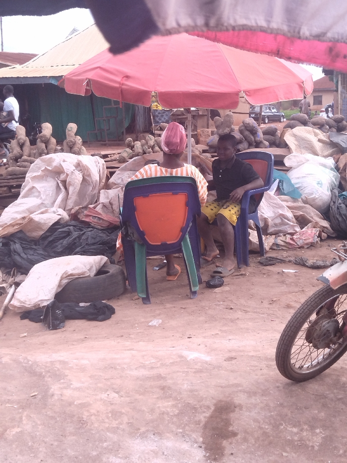 Two people are sitting under a red umbrella. The woman on the left is wearing a yellow and white striped dress and a pink head wrap. The man on the right is wearing a black shirt and yellow and black shorts. They are sitting in plastic chairs. There is a pile of large, brown, round objects behind them. In the background, there are other people, buildings, and a motorcycle. The ground is dirt, and there are many pieces of garbage scattered around. The scene is located in Anyigba, Nigeria, at the coordinates 7.504, 7.178. The location is likely a market area or roadside stall.