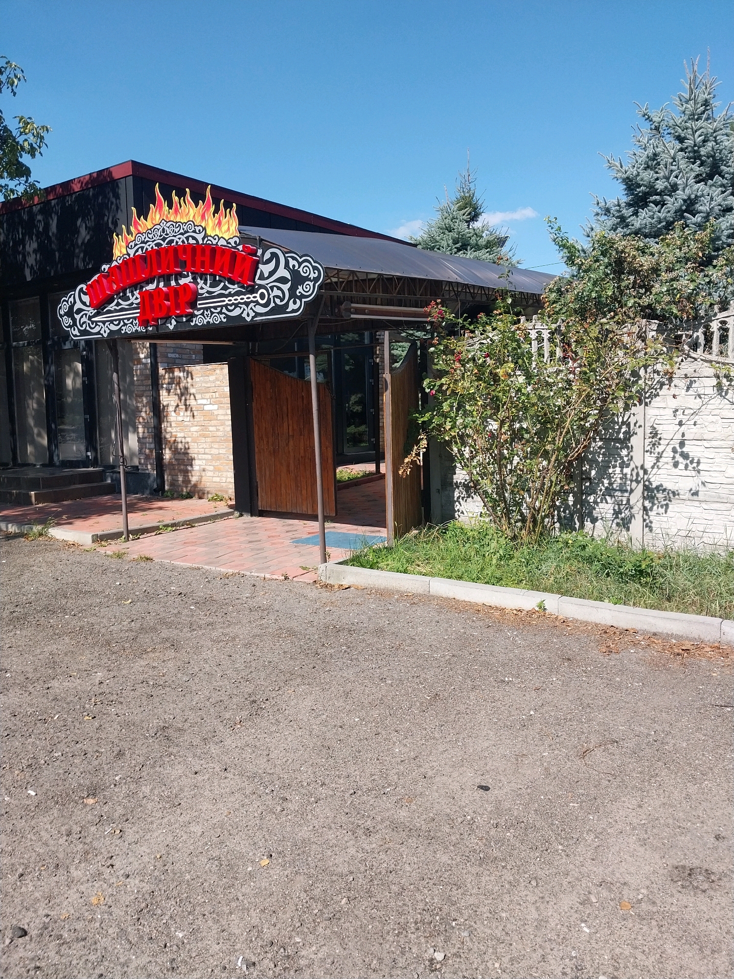The image shows the entrance to a restaurant or cafe with a prominent sign featuring red, flame-shaped lettering. The sign is ornate, with black and white decorative elements. The building has a modern awning over the entrance, which is made of a transparent material supported by a metal frame. The entrance itself has glass doors and a brick facade visible behind the wooden double doors. The area in front of the entrance is paved with reddish-brown brickwork. To the right of the entrance, there is a concrete fence and a lush green bush with some red berries. In the background, several coniferous trees are visible against a clear blue sky. The foreground consists of a gravelly or dirt area. The scene suggests a commercial establishment in an outdoor setting.
This establishment is likely located in Dnipro, Ukraine.