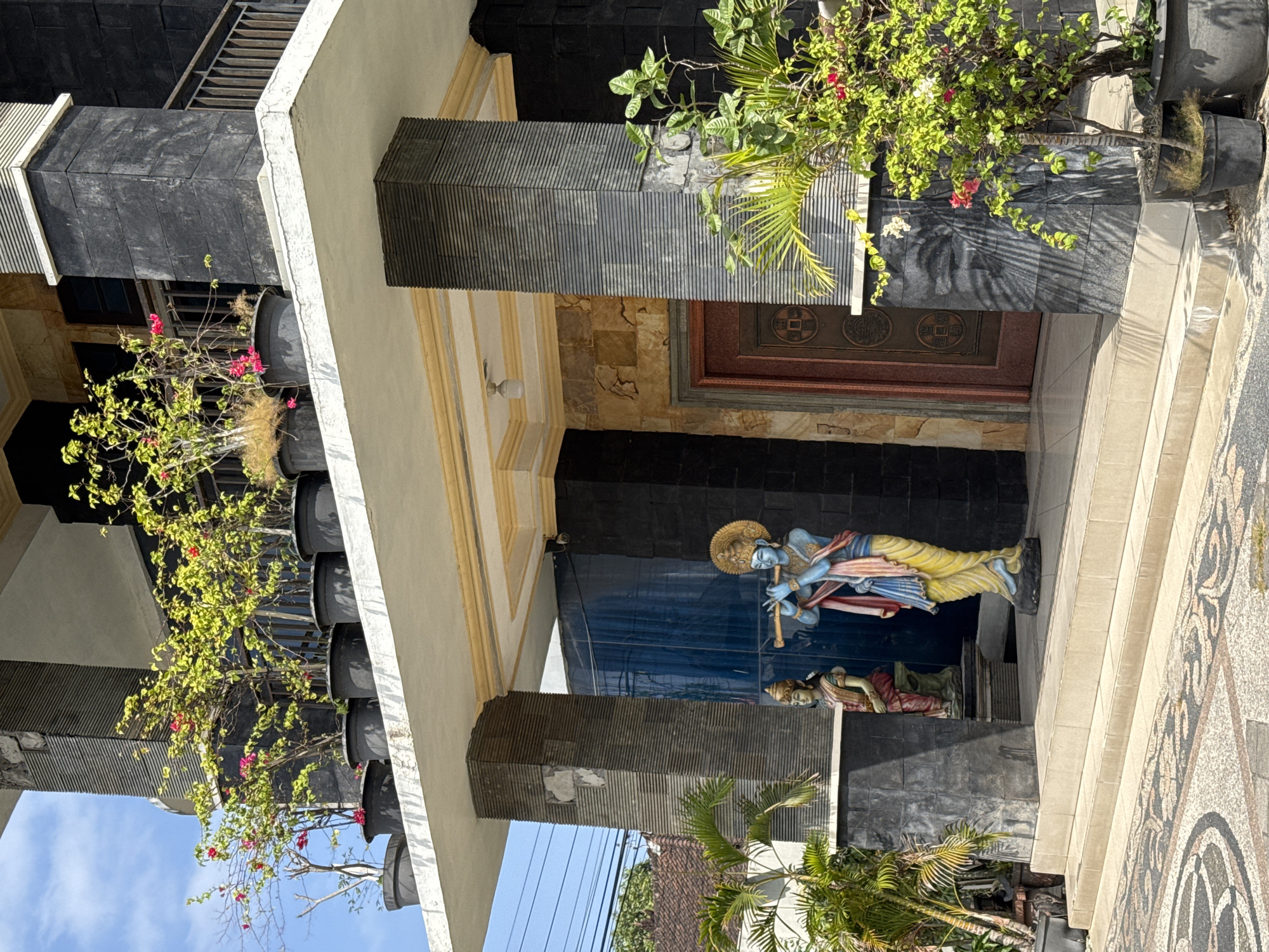 In the city of Denpasar, Indonesia, a scene unfolds in front of a building. The building's facade features black stone accents and a white overhang. On the overhang, several potted plants with green and yellow leaves and pink flowers are visible. In front of the building, two statues stand on a tiled walkway. The statue on the left depicts a seated figure wearing traditional Indian clothing. The statue on the right is of a blue-skinned figure playing a flute, also in traditional attire. These statues are positioned on a raised platform with steps leading up to it. To the right of the statues, a potted plant with green leaves and red and white flowers is situated. The building itself has a large brown door with ornate carvings, flanked by stone pillars. The sky above is a clear blue.