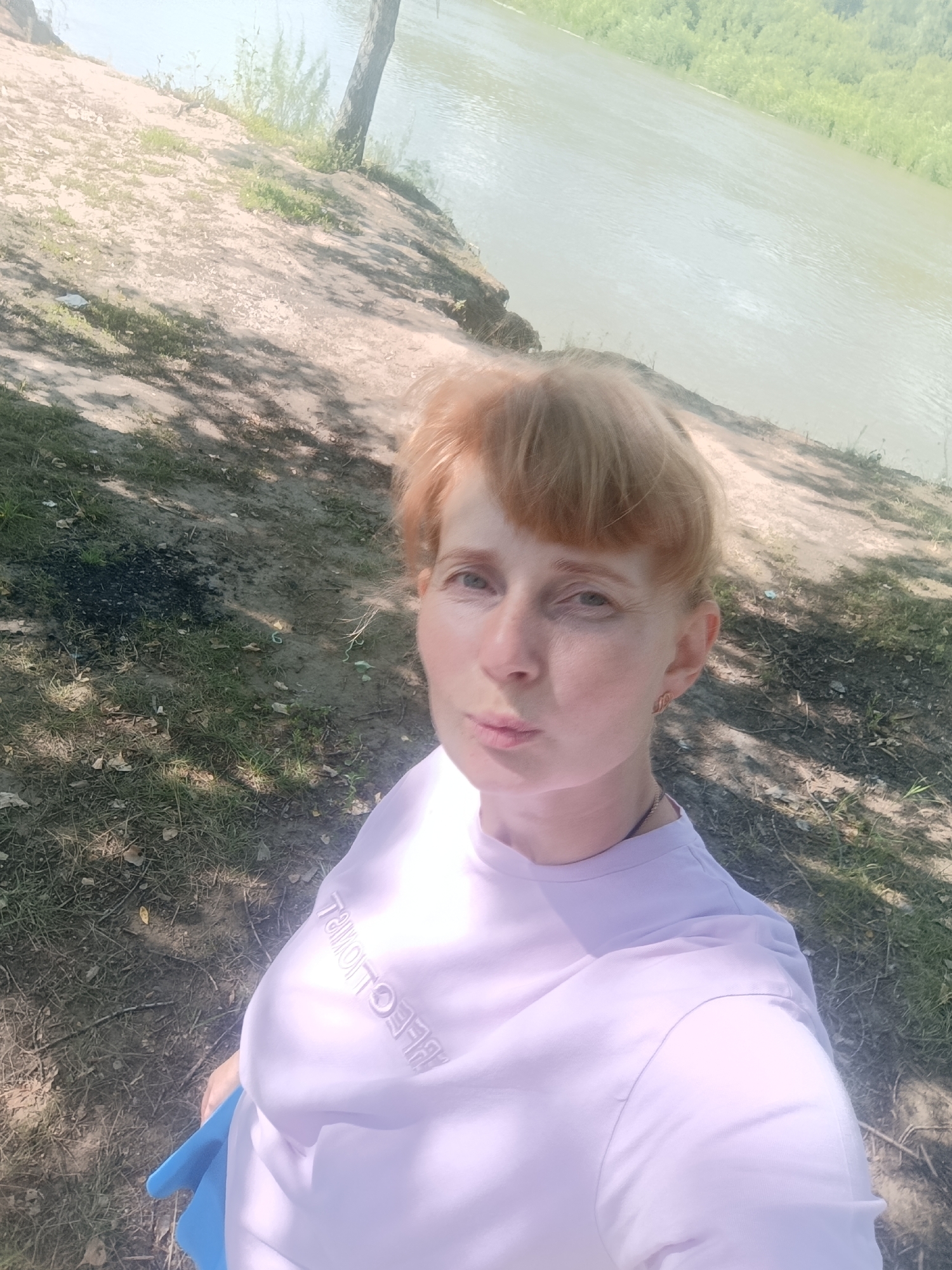 A fair-skinned woman with short, reddish-blonde hair is captured in a selfie outdoors. She is wearing a light purple t-shirt with some text on it, and a blue bag is visible. The woman is positioned on a dirt embankment overlooking a wide, calm river with a treeline on the opposite bank. The ground around her is covered with sparse grass, dirt, and some fallen leaves. Shadows from trees are cast on the ground. The sky is bright and appears to be sunny. The location is identified as Samorodovo, Russia.