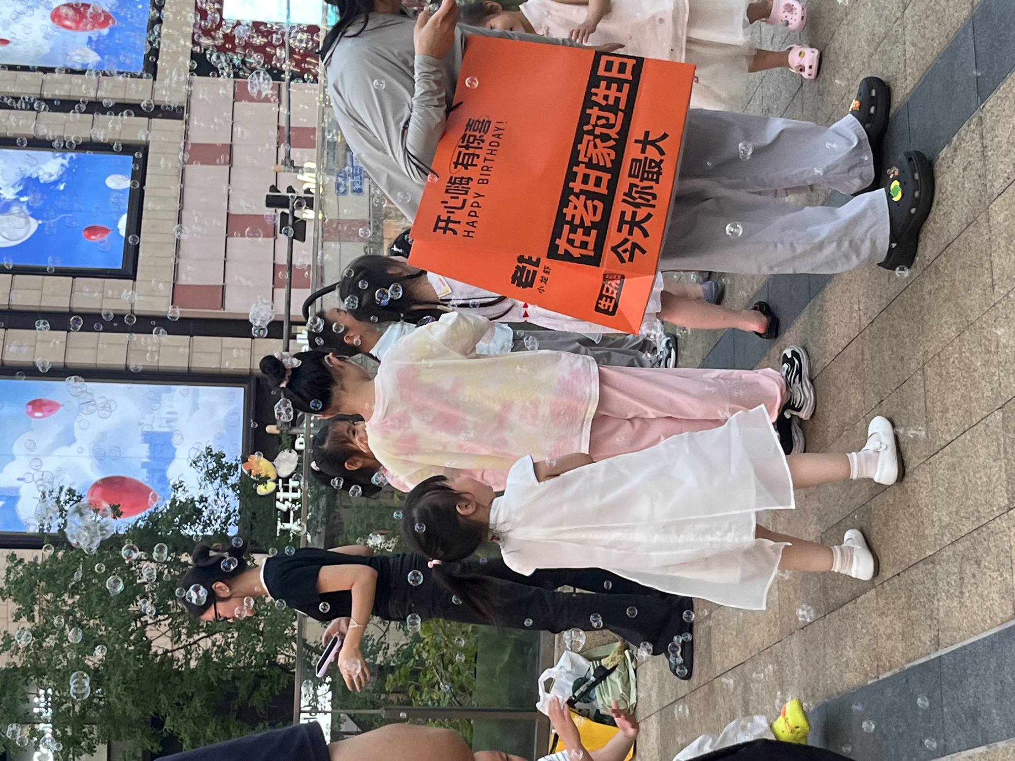 The image captures an outdoor urban scene in Yuzhong District, China, characterized by numerous translucent soap bubbles floating throughout the frame. The setting features a paved pedestrian area with a background of buildings and greenery.
On the left, a woman with dark hair styled in two buns, wearing black-framed glasses, a black short-sleeved top, and black wide-leg pants, holds a pink smartphone in her left hand. Her right hand is cupped, suggesting interaction with an unseen bubble-generating device. Green foliage is visible behind her.
In the center, a group of young children are present. A girl in a white sleeveless dress, white ankle socks, and white flat shoes, with her dark hair in a ponytail, stands facing right, looking upwards towards the bubbles. Another child wears a yellow, pink, and white tie-dye t-shirt with pink pants, facing away from the viewer.
On the right, an adult figure, wearing a light grey long-sleeved shirt and grey pants, is bent over, holding a prominent orange shopping bag. This individual wears black Crocs adorned with colorful charms. The orange bag displays "HAPPY BIRTHDAY!" in white English text, along with Chinese characters reading "开心嘛 有惊喜" (Happy, there's a surprise), "在老甘家过生日" (Celebrating birthday at Old Gan's house), and "今天你最大" (Today you are the biggest/most important). Other children, including one in a light pink top and pink Crocs, are partially visible on the right.
The background features large digital display screens integrated into a building facade, showing an image of a blue sky with white clouds and floating red and white balloons. The ground consists of tan and grey rectangular pavers. A yellow bag and a white plastic bag are on the ground on the far left.
