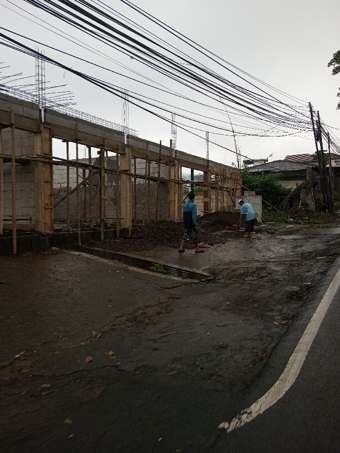 The image depicts an outdoor scene in the city of YonPhoogla, Bhutan, featuring a construction site alongside a wet asphalt road under an overcast sky.
In the foreground, a dark asphalt road, damp and containing scattered debris, runs from the lower left to the right, with a solid white line marking its right edge. A metal drain grate is visible on the left side of the road.
To the left, a single-story structure is under construction. It consists of light-colored block walls, concrete columns, and a concrete beam running horizontally across the top. Wooden scaffolding and temporary supports are integrated into the structure. Exposed rebar protrudes vertically from the tops of several columns. Piles of dark aggregate and loose soil are present on the unpaved ground directly in front of the construction.
Two adult males are visible near the construction. The individual on the left wears a blue long-sleeved shirt and patterned shorts, standing barefoot with his back partially turned. The individual on the right wears a light blue t-shirt and dark shorts, also barefoot, bending over near a small stack of light-colored bricks or blocks, appearing to be engaged in work.
Numerous black utility lines traverse the upper portion of the frame, strung overhead and connecting to a utility pole on the far right. In the background, behind the construction site, residential buildings with tiled roofs are visible amidst green foliage.
