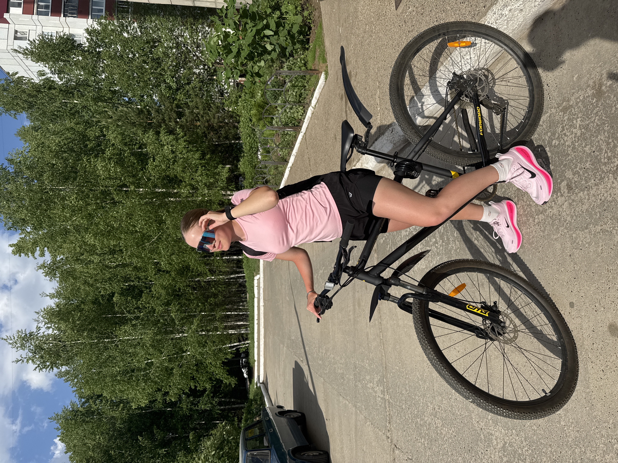 A young woman stands next to a black mountain bicycle on a paved surface in Chaikovsky, Russia. She wears a pink t-shirt, black shorts, white socks, and bright pink sneakers. Sunglasses are perched on her head. The bicycle is lying horizontally on the pavement, partially obscuring the woman's legs. Behind her are tall trees and a building. An older model car is partially visible in the lower left corner. The setting appears to be an urban or suburban street with green space.