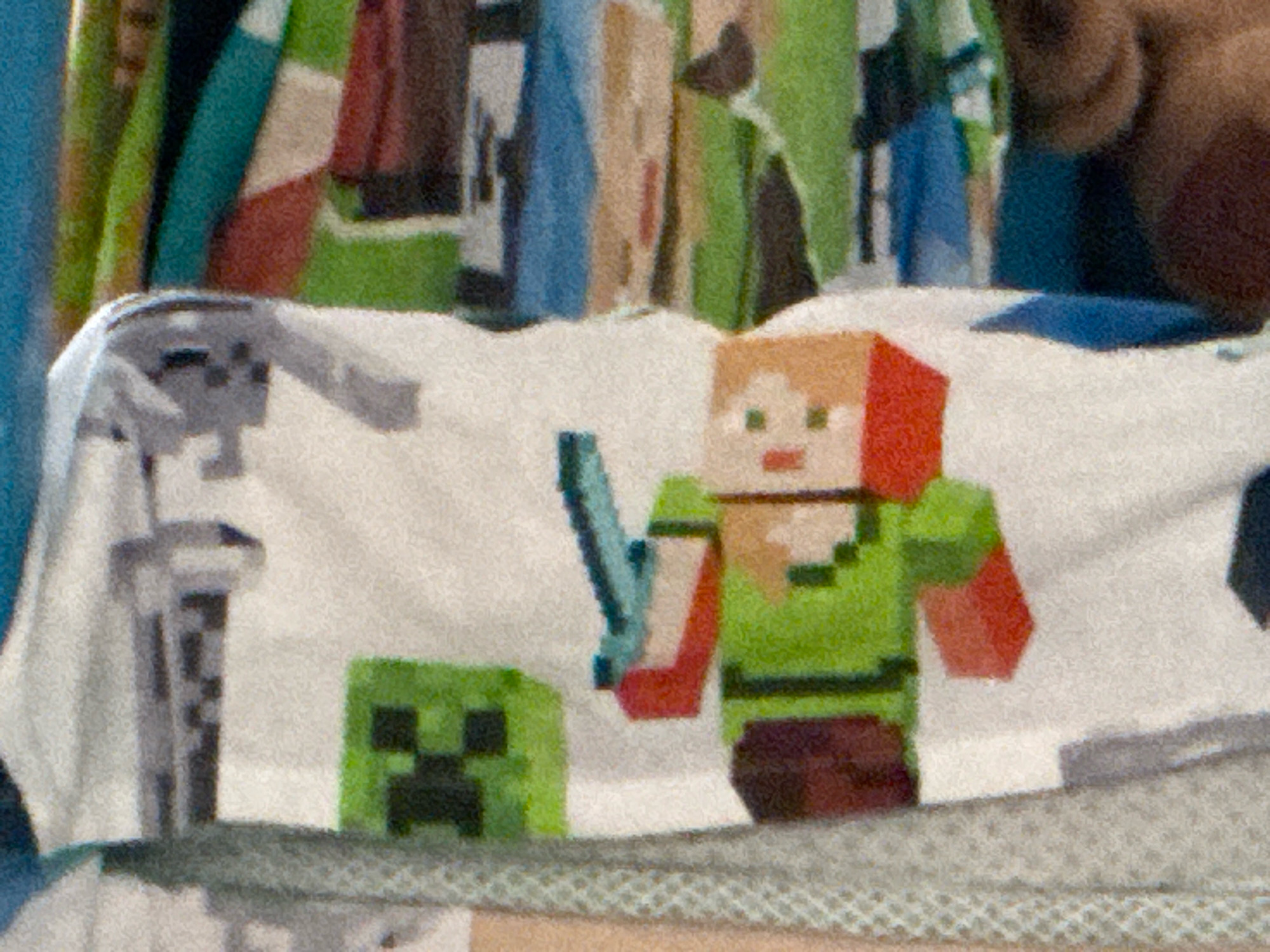 The image shows a close-up of a white fabric with pixelated graphics of Minecraft characters. On the left is a green creeper character with a black face. To its right is a pixelated representation of Alex, a female Minecraft character, wearing a green top and brown shorts, holding a light blue sword. Behind the characters, the fabric appears to have more colorful, abstract pixelated patterns. The image is grainy, suggesting it was taken with low light or a camera with a high ISO setting. The context of the image and its location in Netanya, Israel, is not discernible from the visual information provided.
