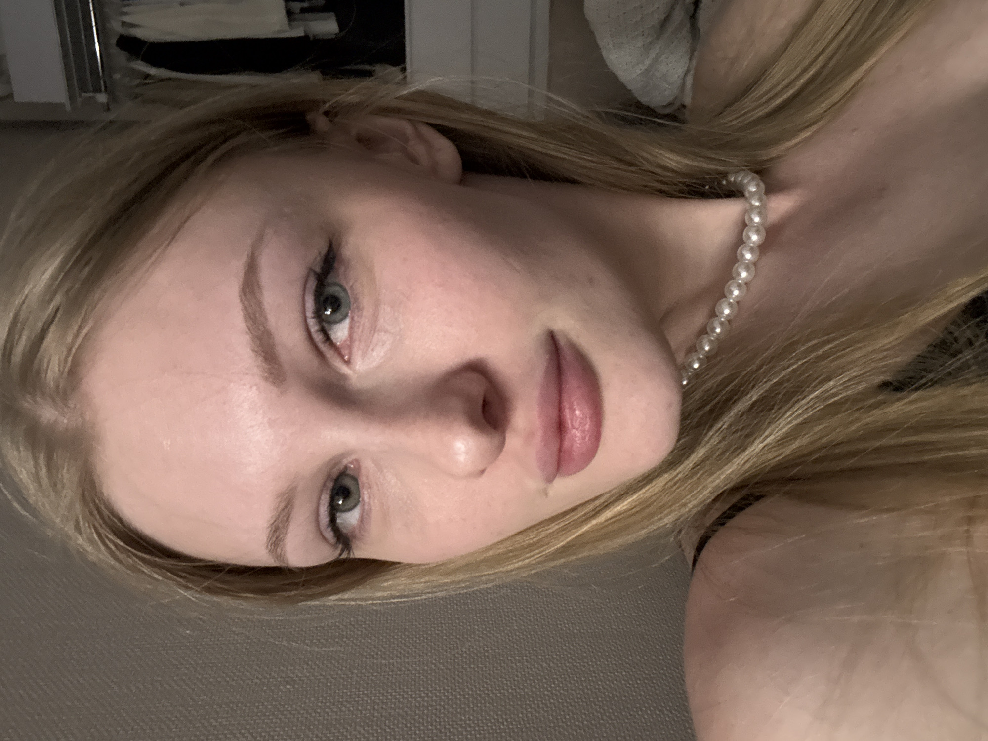 A young woman with blonde hair, blue eyes, and fair skin is shown in a close-up, selfie-style portrait. She is wearing a pearl necklace. The background appears to be indoors, with a textured wall on the left and clothing on hangers visible on the right. The lighting is soft and appears to be from a lamp or natural light source. There are no visible indications of the city of Perm, Russia, or any specific environmental context that would allow for its identification.