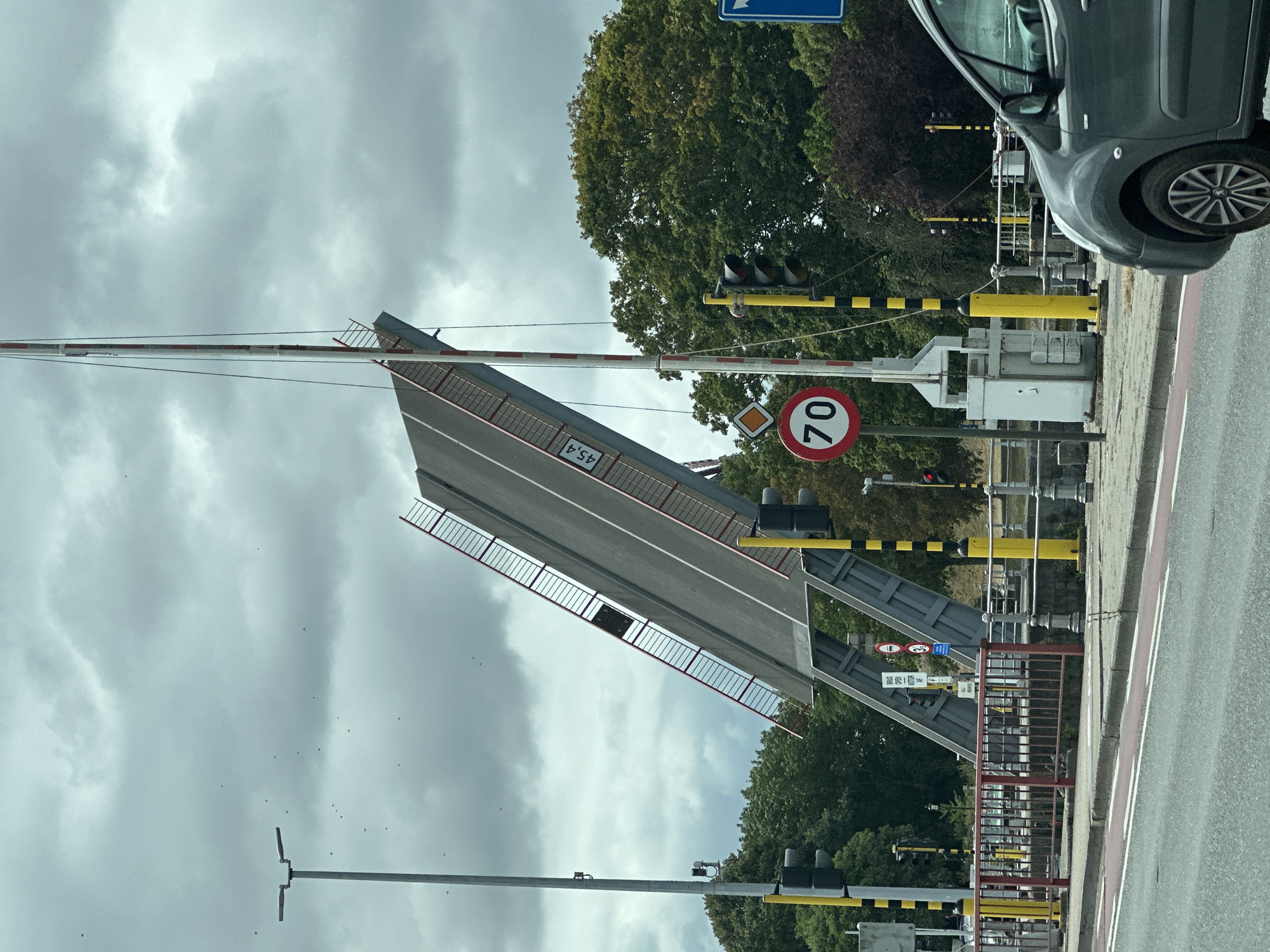 This is a photograph of a bascule bridge in Bruges, Belgium, in an open position, allowing passage for boats or ships. The bridge is raised at a steep angle, with a section of the roadway extending upwards. The sky is overcast with dark, heavy clouds, and there are several small, indistinct objects that appear to be birds or debris scattered across it.
In the foreground, a grey car is visible on the right side of the frame, partially obscuring the view of the bridge's base. A red and white striped pole, part of the bridge's warning system, is positioned vertically to the right of the raised bridge section. Traffic lights, one red and one showing green, are visible on poles on both sides of the roadway leading up to the bridge. A circular speed limit sign with "70" is posted on the left side.
Yellow and black striped poles, typical of traffic control in Belgium, are also present. The bridge itself has red railings along its elevated section, and a sign with "45,4" is visible on its side. The background consists of lush green trees and some darker foliage, suggesting an urban or park-like setting. The overall impression is a functional piece of infrastructure in use, with a hint of dramatic weather.
