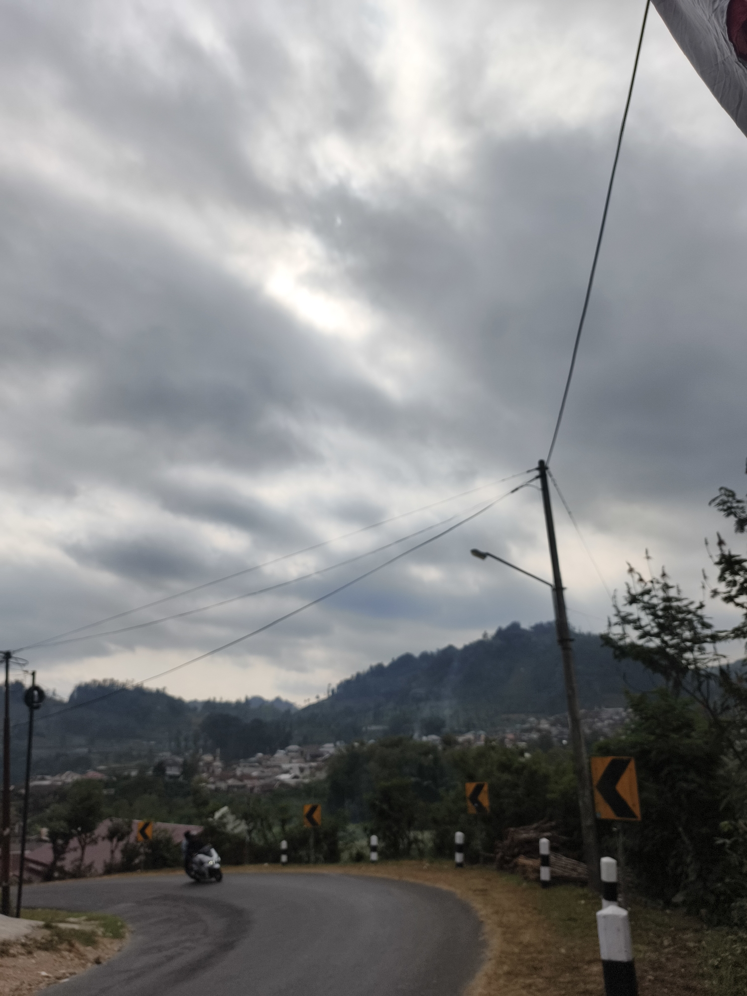 The image depicts a winding road in a hilly, semi-rural area. The sky is overcast with patches of sunlight breaking through. A motorcycle is visible on the road, turning a curve. Road signs indicating curves are present. Utility poles and wires crisscross the scene. In the background, a village or town is nestled among the hills, with buildings and trees. The overall impression is a somewhat hazy, natural landscape with elements of human infrastructure.