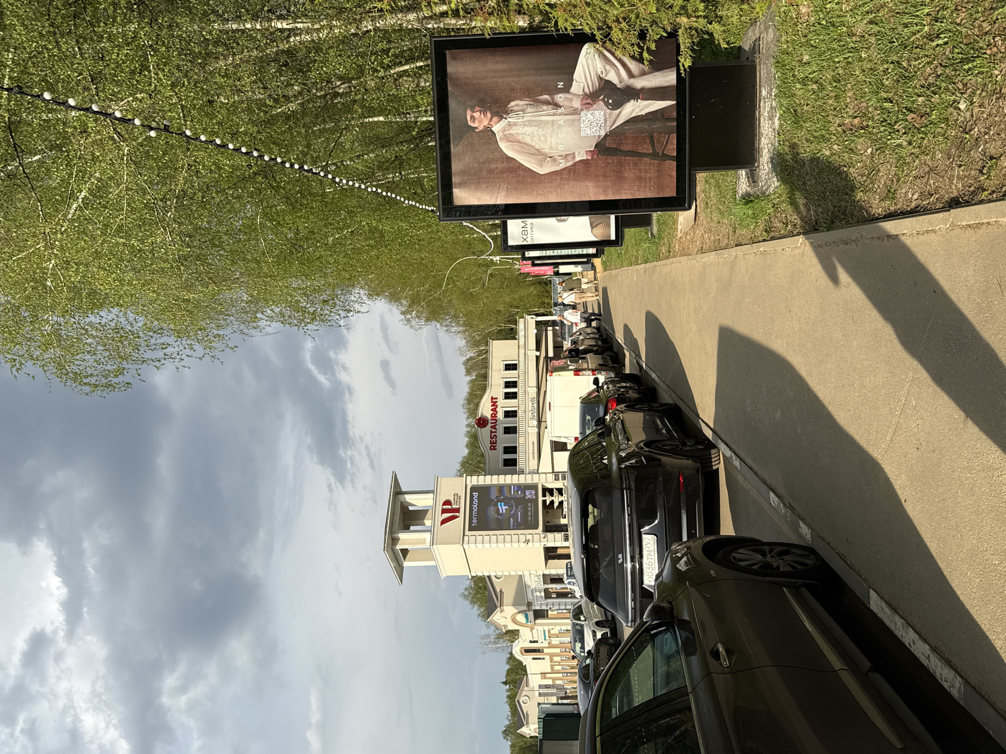 The image depicts a street scene in Vnukovo, Russia. The view is along a sidewalk lined with advertising billboards. Adjacent to the sidewalk is a row of parked cars. Buildings are visible in the background, including one with "RESTAURANT" in red letters and another with the "VP termoland" logo and a digital screen. A string of lights is strung among the trees on the left side of the image. The sky is overcast with clouds. The license plate visible on one of the cars appears to read "O936TM797".