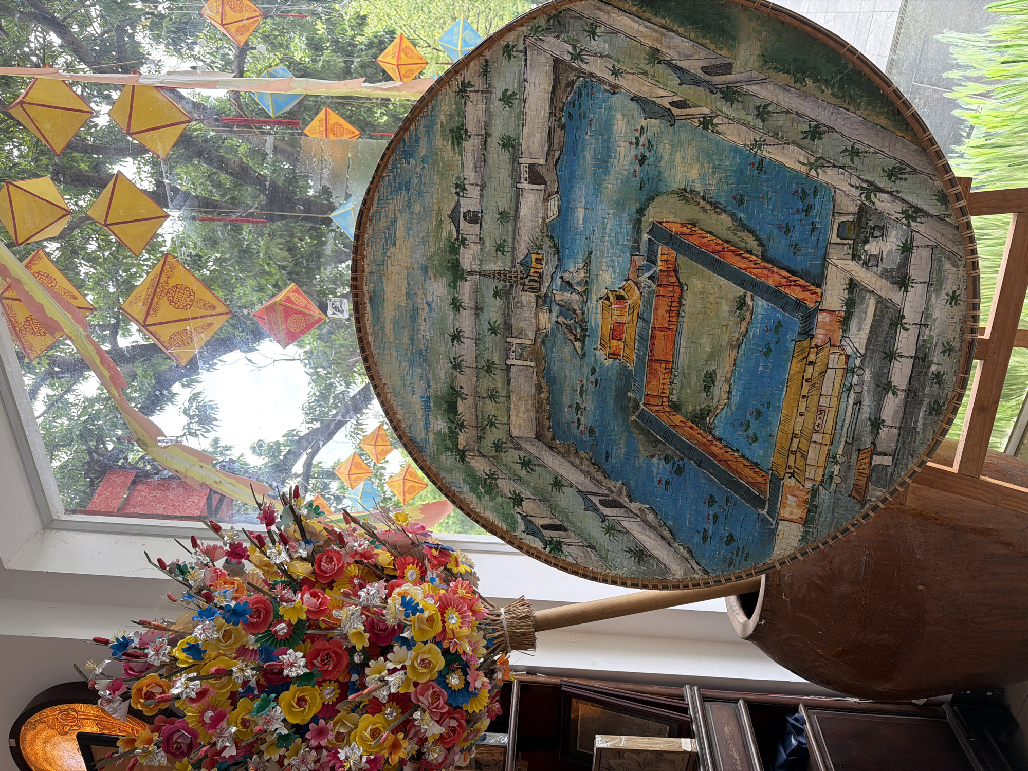 A decorative, woven fan with a painted scene of a traditional Vietnamese palace and garden is prominently displayed. The fan depicts a courtyard with a central pond, ornate buildings with orange and yellow roofs, pagodas, and lush greenery, all rendered in a somewhat stylized manner. Behind the fan, a large, rustic brown ceramic pot holds a vibrant bouquet of artificial flowers in various bright colors. A wooden handle extends from the bouquet and rests inside the pot. In the background, through a window, numerous colorful diamond-shaped lanterns, predominantly yellow with red accents bearing the inscription "HUE," hang suspended, indicating an outdoor setting. The overall impression is one of cultural display and festive decoration, potentially at an event or establishment in Thuận Hóa Ward, Vietnam, given the mention of the city on the lanterns.