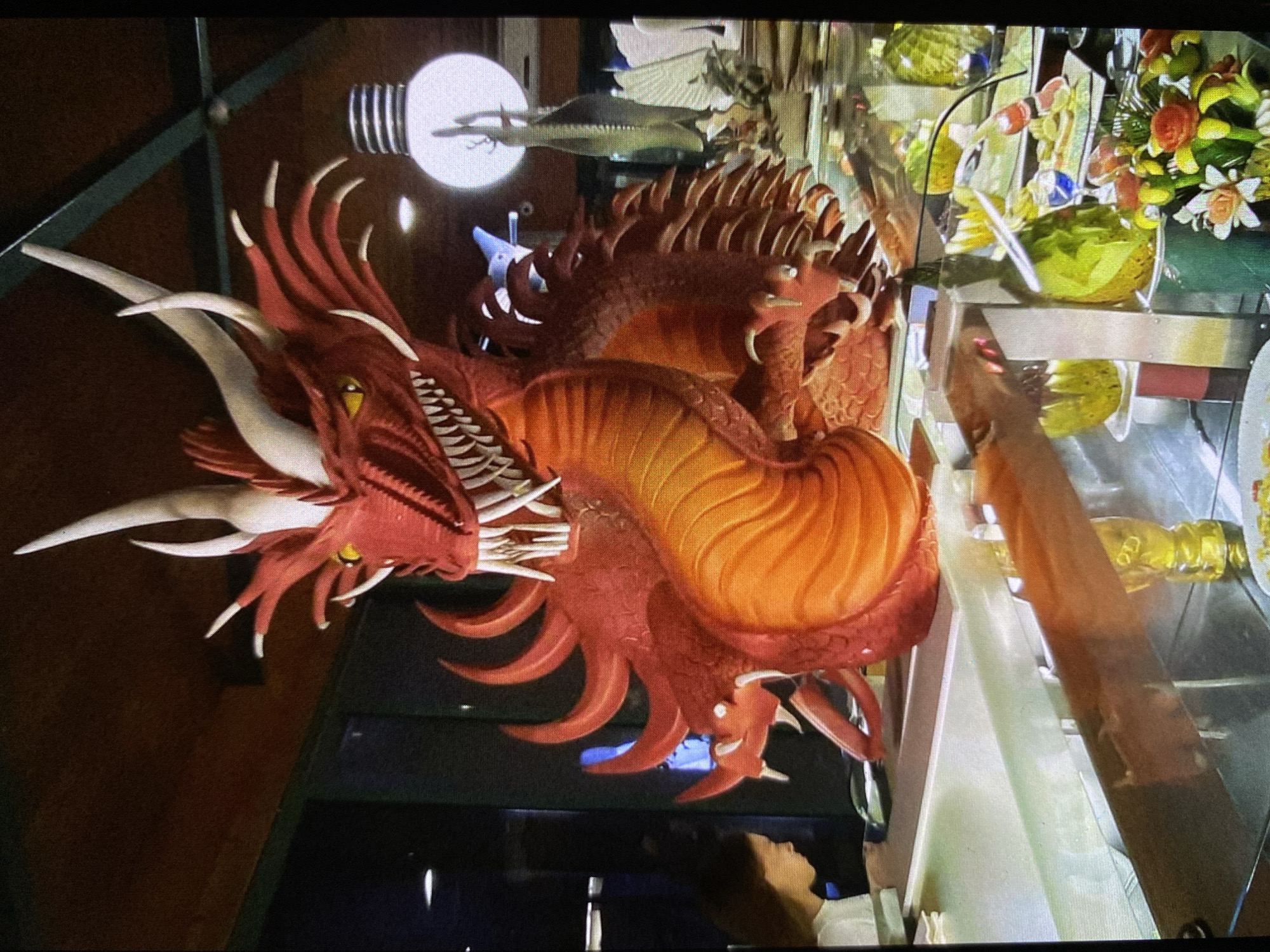 The image displays a vibrant, intricately crafted red and orange dragon sculpture, which serves as the central focal point. The dragon features white horns, sharp white teeth, and bright yellow eyes, giving it a fierce yet artistic appearance. Its body is detailed with scales and a distinct, ribbed orange underbelly, suggesting it might be a food carving, such as from fruit or sugar, or a decorative centerpiece.
The dragon is positioned in front of, or within, a display case or counter, which is made of glass and contains various items. Behind the dragon's head, through the glass, a large, white, lightbulb-shaped decoration is visible. Further back, on shelves within the display, are dishes containing green and yellow items, possibly salads, fruits, or desserts. There are also floral arrangements with yellow and orange flowers, and bottles that appear to hold liquids like oil or dressing.
The scene suggests an indoor setting, likely a restaurant, hotel buffet, or a catering event, given the elaborate food display and decorations. The lighting appears to be artificial, typical of an indoor venue. Reflections are visible on the glass surfaces, including a partial reflection of a person's head and shoulders in the lower right corner, indicating human presence. There is no visible text in the image. This elaborate display is located in Bankya, Bulgaria.