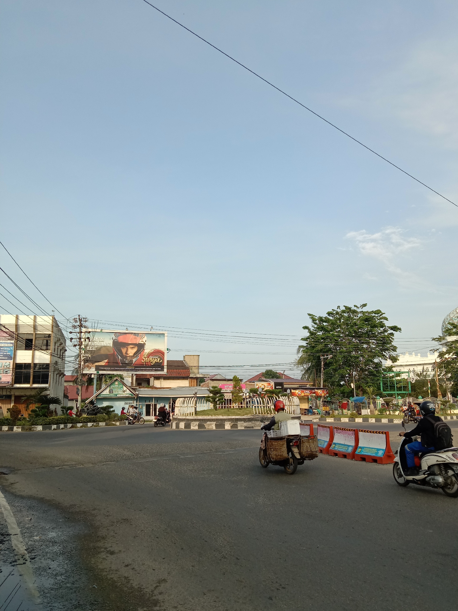 The image depicts a street scene in Lhokseumawe, Indonesia. The sky is pale blue with thin cloud cover. In the foreground, there is a gray asphalt road. A person is riding a motor scooter in front of a line of orange and white temporary road barricades. A delivery man is riding a motor scooter with a crate of goods. In the background, there is a roundabout in the center of the intersection with a lawn and plants. Buildings surround the roundabout, with the skyline including a building with a dome and a large billboard advertising a brand of cigarettes. Overhead powerlines are visible. Several vehicles are driving through the roundabout. The scene appears to be taken during daylight.