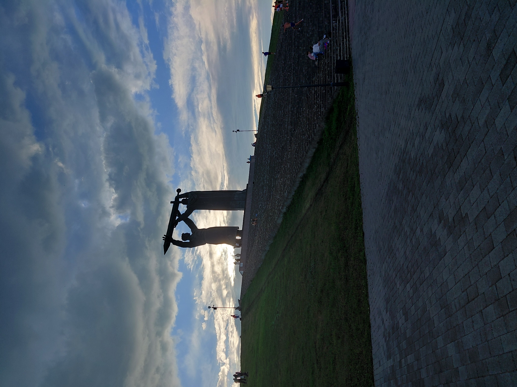 The image depicts the Monument to the Magnitogorsk Steelworkers in Magnitogorsk, Russia, set against a dramatic sky with clouds. The monument, a silhouette against the setting sun, features two figures holding a sword aloft. A grand staircase leads up to the monument, with several people visible ascending and sitting on the steps. To the left of the stairs is a grassy slope, where more people are gathered. The foreground is a paved area. The lighting suggests it is late afternoon or early evening.