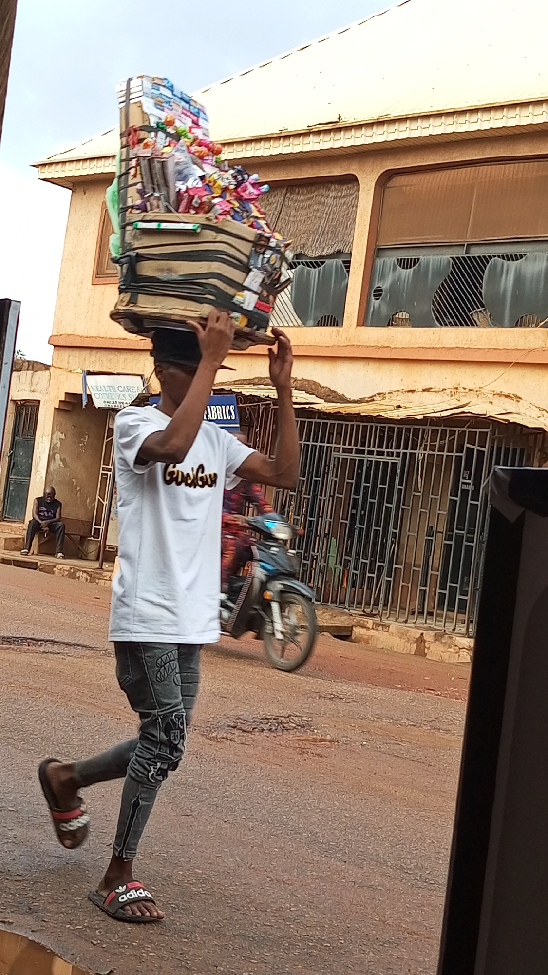 A man in Sabon Tasha, Nigeria, is carrying a large, rectangular stack of goods on his head. The items are secured with tape and string and appear to be various small goods, possibly for sale. The man is wearing a white t-shirt, grey patterned jeans, and Adidas slides. He is walking on a street, and a motorcycle is partially visible in the background. In the background there are buildings. One has a metal grate door, and what appear to be damaged windows. Another person is sitting on a step nearby.