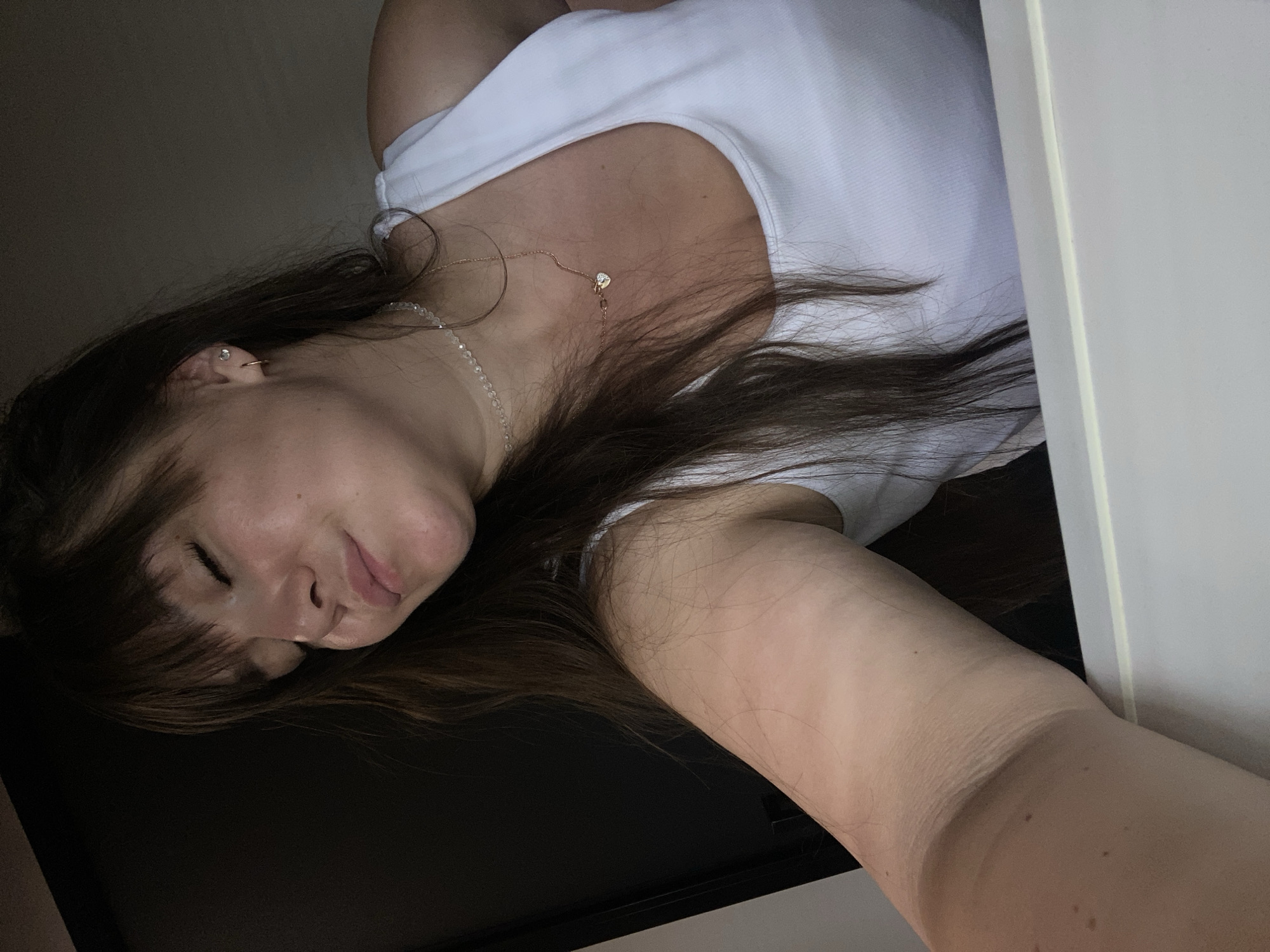 A young woman with long brown hair and bangs is taking a selfie in what appears to be a dimly lit indoor setting. She is wearing a white tank top and a delicate necklace. Her eyes are closed, and she has a relaxed expression. A subtle tan line is visible on her left arm. The background is dark, with a hint of a door or wall on the left and a lighter surface, possibly a table or desk, in the foreground. There is no discernible information to place this image in Votkinsk, Russia.