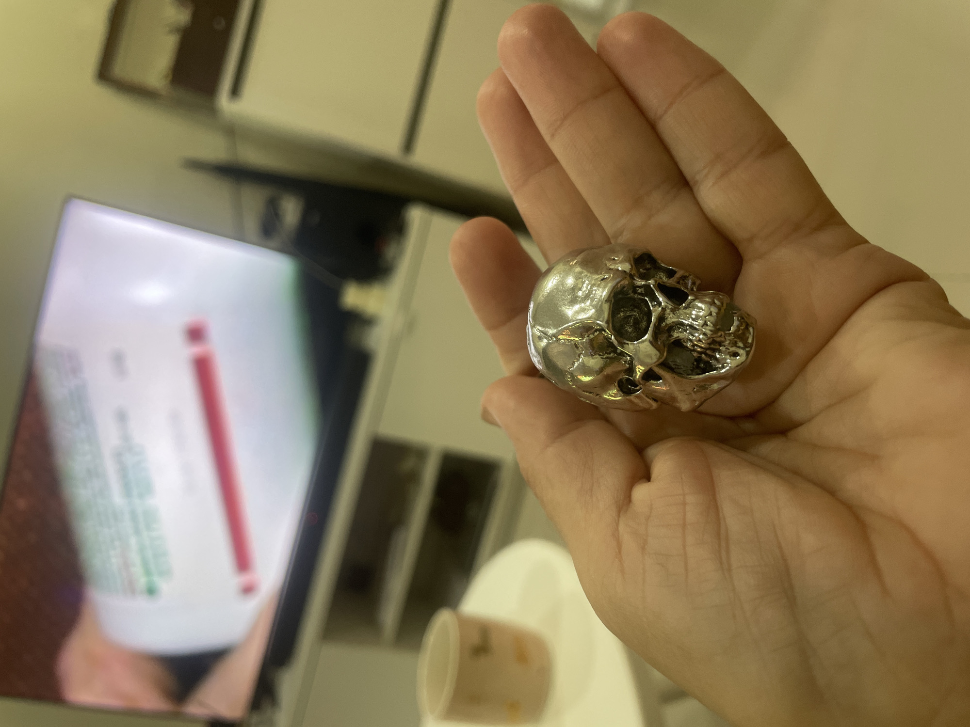 A person holds a small, shiny, silver-colored skull in their palm. The skull is detailed and appears to be made of metal. In the background, out of focus, there is a computer monitor displaying an image of a bottle with a red stripe. Also visible are parts of furniture, including cabinets and a cup on a white surface. There is no definitive visual indication of the location being in Thep Krasatti, Thailand, or any specific city. The scene is indoors and likely a personal space.