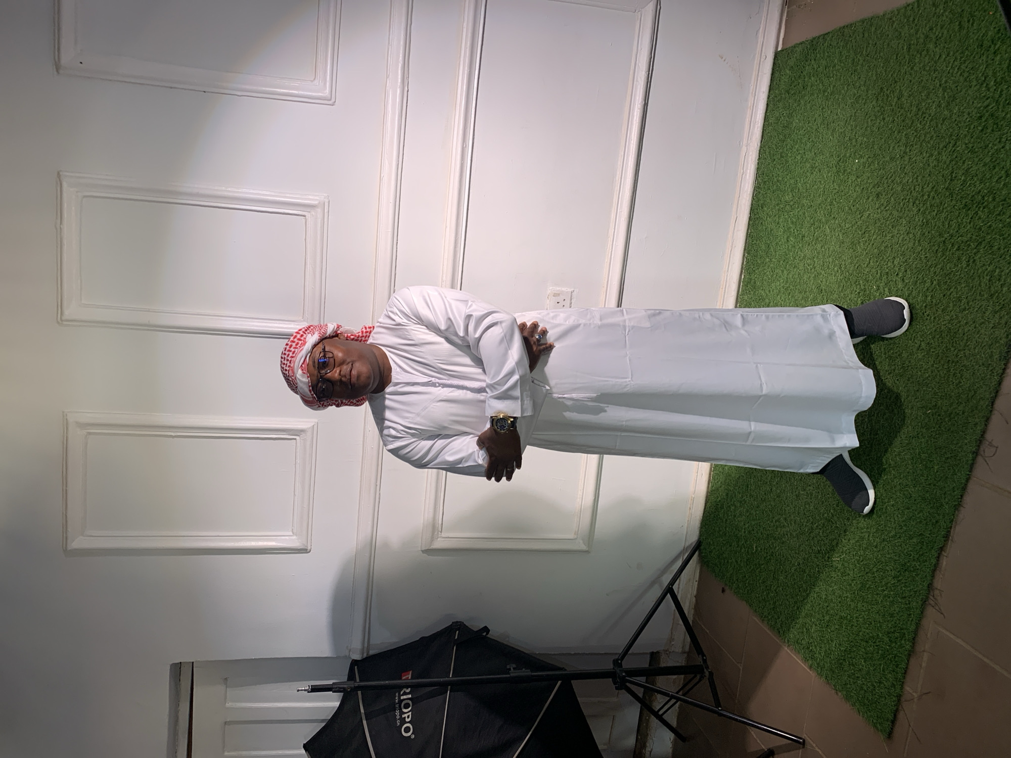 A man stands indoors, likely in a photography studio. He wears traditional Middle Eastern attire: a white thobe and a red and white checkered keffiyeh. He also wears glasses and a watch. He has his arms crossed. The background consists of a white door or wall paneling with rectangular indentations. There's a patch of green artificial turf to the right. A photography softbox with a tripod stand is visible near the bottom of the frame. The location is not specified in the image, and therefore cannot be identified. The image does not contain location metadata to determine its location in Jos, Nigeria, or elsewhere.