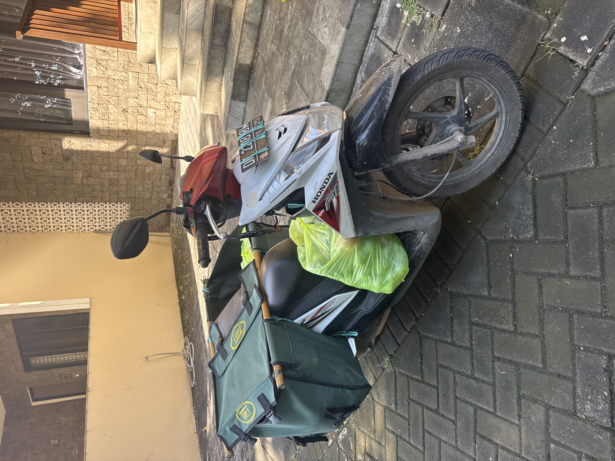 A white and red Honda motorcycle with a green delivery bag attached to the back is parked on a brick-paved area in Kuta Selatan, Indonesia. A license plate is affixed to the front, held on with green rubber bands. A yellow plastic bag is placed on the seat. The motorcycle is parked near a building with a stone exterior and a staircase leading up.