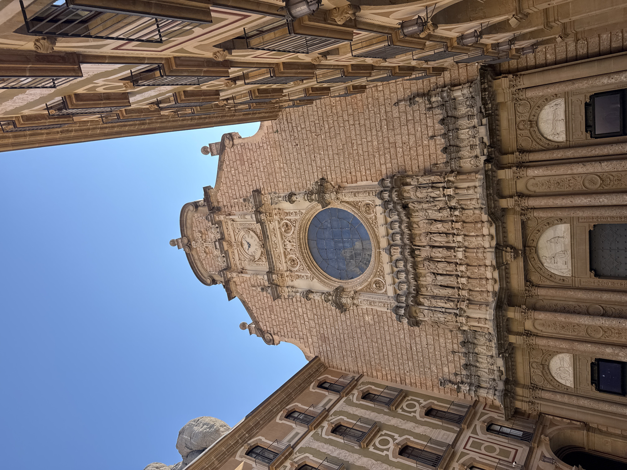 The image shows the facade of the Montserrat Abbey in Monistrol de Montserrat, Spain. The architecture is predominantly Romanesque with Gothic influences, characterized by its stone construction and ornate detailing. The main facade features a large rose window, sculptures of religious figures, and intricate carvings. The building is flanked by other structures, one of which appears to be residential, with balconies and arched windows. The sky is clear and blue, suggesting a sunny day. No people are visible in the image, and no specific actions or interactions are depicted. The scene is static and focuses on the architectural grandeur of the abbey and its surroundings.