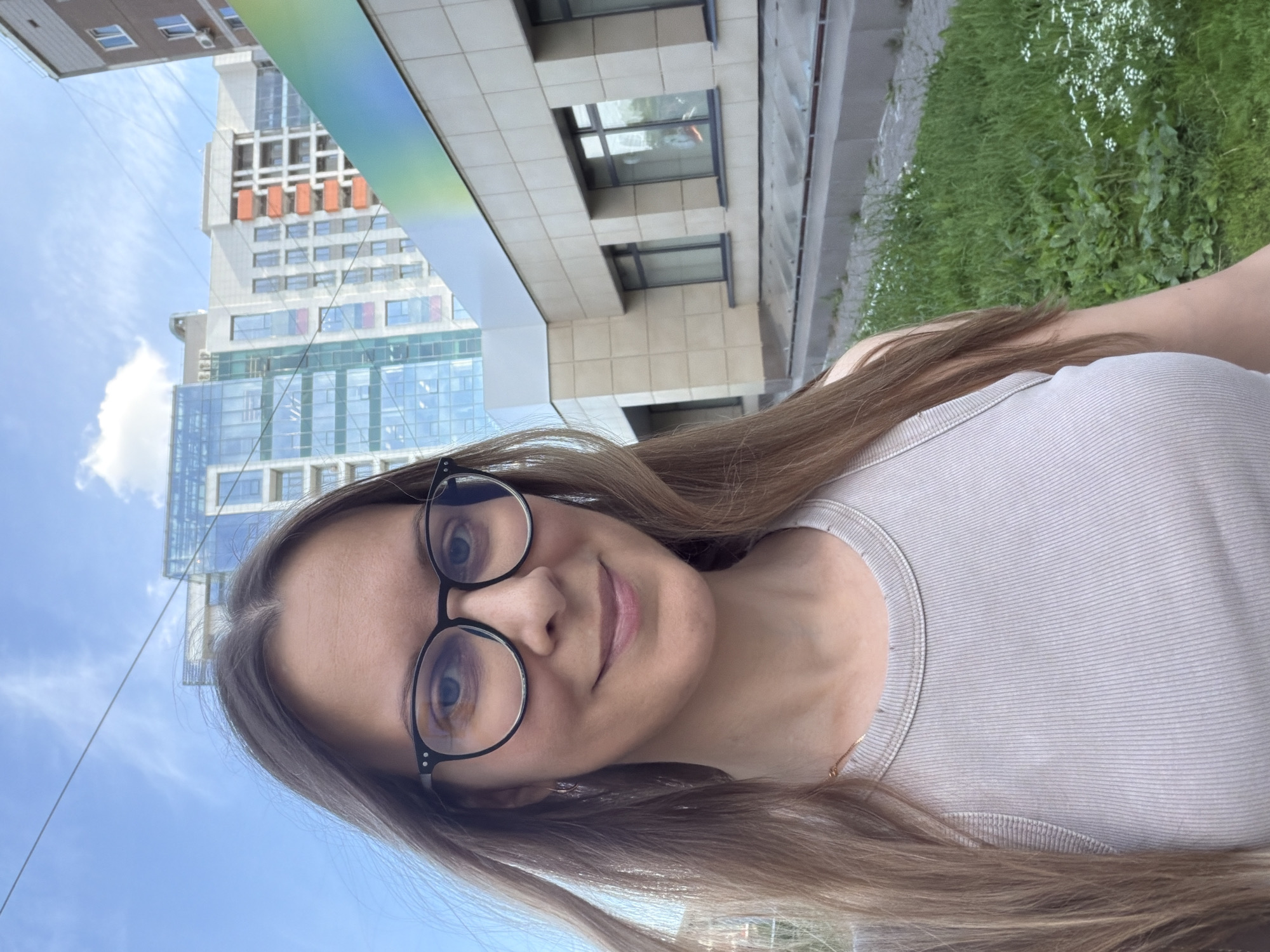 A young woman with long, light brown hair and glasses is taking a selfie outdoors. She is wearing a light-colored, ribbed tank top. Her eyes are blue, and she has a slight smile. In the background, a modern building with glass windows and orange accents is visible, along with a patch of green grass and flowers. The sky is blue with some white clouds. The image appears to be taken on a sunny day, possibly in the afternoon. The overall mood is casual and relaxed. The location is identified as Izhevsk, Russia. There is no visible text in the image.