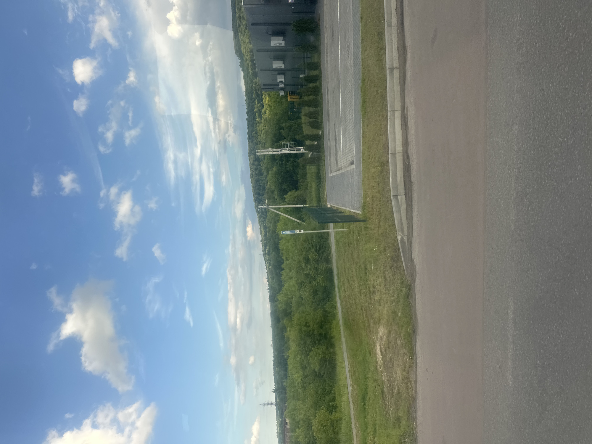 The image captures a wide landscape view under a bright blue sky with scattered white clouds, suggesting a sunny day. In the foreground, a paved road runs horizontally across the bottom of the frame. Beyond the road, a strip of green grass is visible, bordered by a concrete curb.
Further back, a grey asphalt parking lot with white lines marking parking spaces is situated. A dark grey building with a modern architectural design, featuring several air conditioning units mounted on its exterior, stands to the right of the parking lot. In front of the building, a row of small evergreen trees is planted. To the left of the parking lot, a tall white utility pole with equipment attached is visible, and a green metal fence encloses the parking area and a small section of grass. A blue and white parking sign is attached to a pole within the fenced area.
The background is dominated by a lush, green, rolling landscape of dense trees and vegetation, forming a wooded hillside. In the distance, on the left side of the frame, a tall lattice tower, possibly a power line pylon, is visible above the trees. The overall scene suggests an industrial or commercial area bordering a natural, hilly environment. The location is identified as Zolochiv, Ukraine.