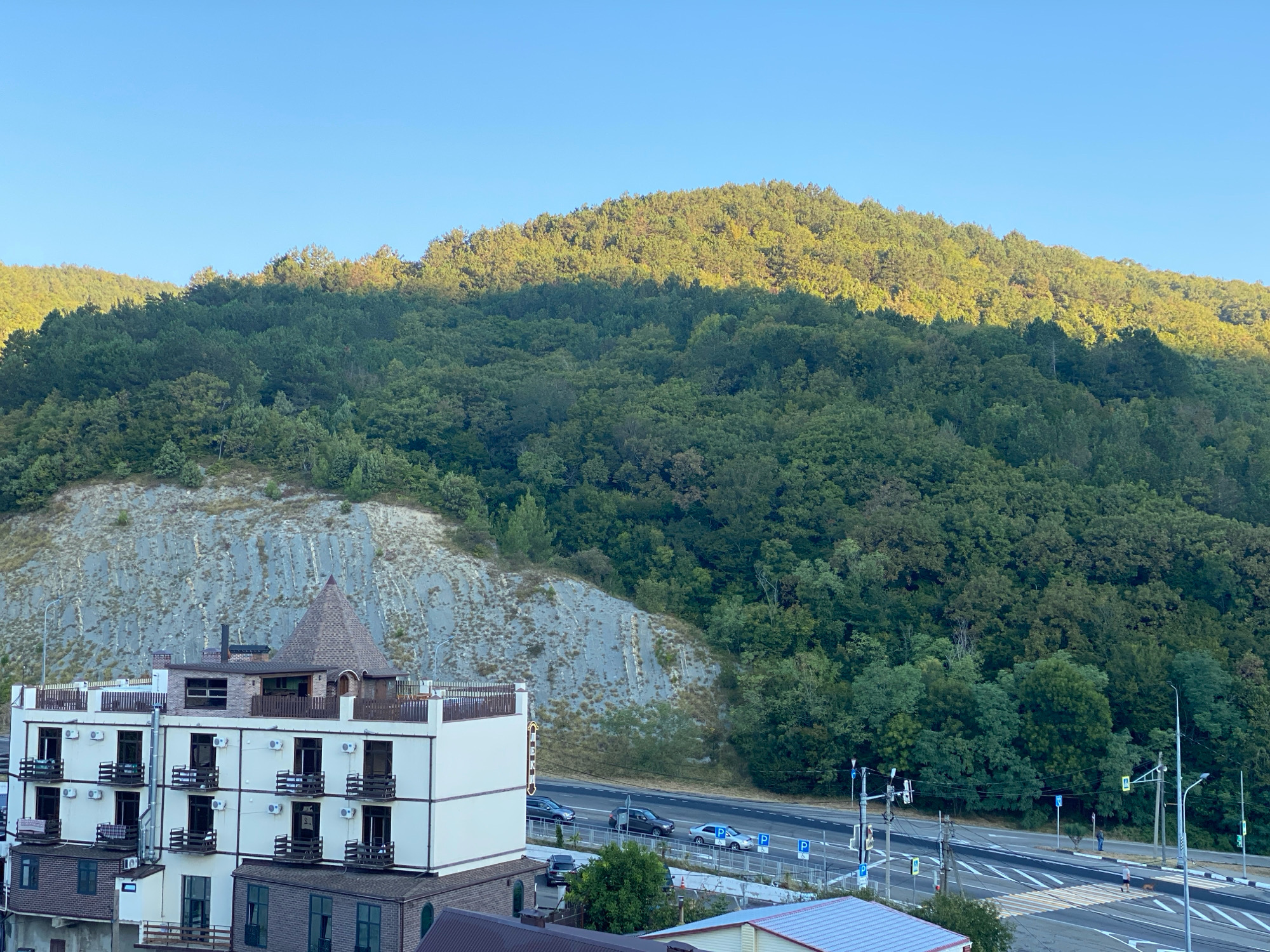 The image depicts a scene in Nebug, Russia, with a multi-story building in the foreground and a densely wooded hillside rising behind it. The building, painted white with brown accents, features multiple balconies with dark railings and air conditioning units. At the top of the building, a conical structure with a brown roof is visible.
To the right of the building, a road with multiple lanes is visible, with several cars driving on it. Streetlights and traffic signals line the road. Pedestrians can be seen crossing the road at a marked crosswalk. Parking signs are also present.
The hillside behind the building is covered in a thick forest of green trees. A portion of the hillside, to the left of the trees, is a sheer rock face with visible striations. The sky above is clear and blue, with the upper slopes of the hill illuminated by sunlight, casting shadows on the lower parts. The overall impression is of a developed area nestled against a natural landscape.