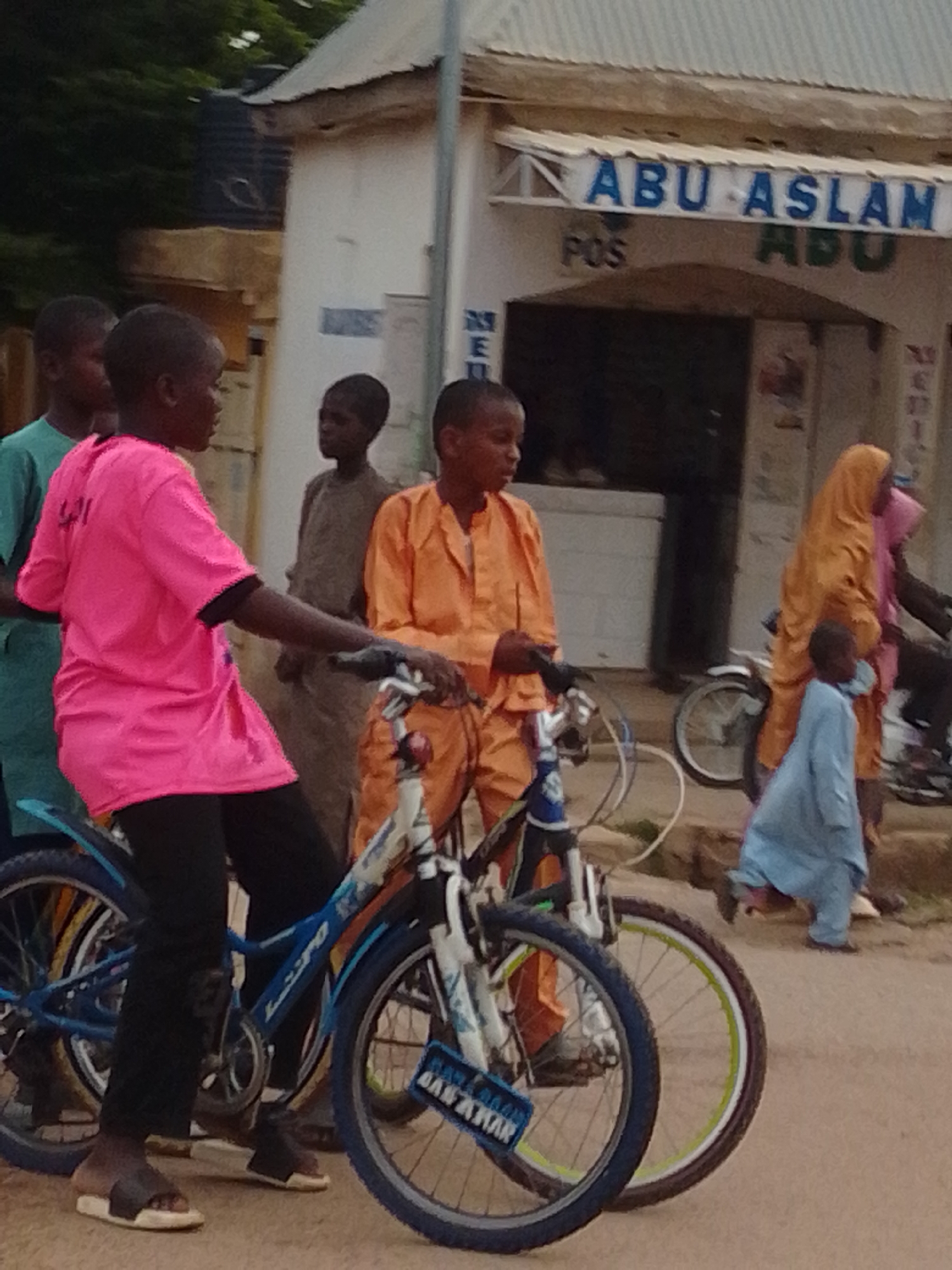 The image depicts a street scene in Gombe, Nigeria, with several individuals and bicycles. In the foreground, a person in a vibrant pink t-shirt and black pants is standing next to a blue bicycle. Another person in an orange outfit is partially visible behind them, also near a bicycle. To the left, a person in a green t-shirt is standing, and further back, a person in a brown garment is visible. On the right side of the image, a person in a yellow hijab and a child in a blue garment are present, with a bicycle behind them. The background features a building with signage that reads "ABU ASLAM POS ABU." The overall environment appears to be an outdoor marketplace or street, with the ground covered in sand or dirt. The lighting suggests it is daytime.