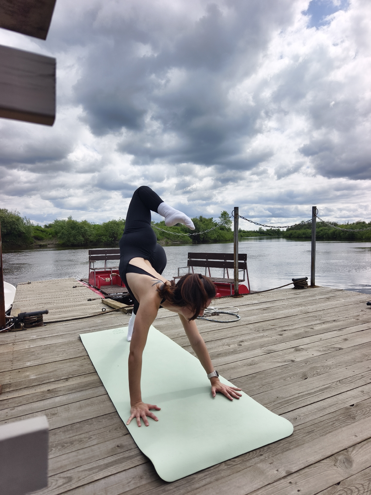 A woman is performing a yoga pose on a wooden pier. She is on a light green yoga mat, with her body in a balanced position. Her left leg is extended upwards and bent at the knee, while her right leg is bent with her foot on the mat. Her arms are extended, with her right hand on the mat and her left arm reaching up. She is wearing black yoga pants and a black sports bra with white socks.
The pier is made of wooden planks and is situated next to a body of water. In the background, there are trees and a cloudy sky. There are also two benches and a red structure on the pier. The scene appears to be outdoors, possibly in a natural setting. The location is in the city of Онохино, Russia.