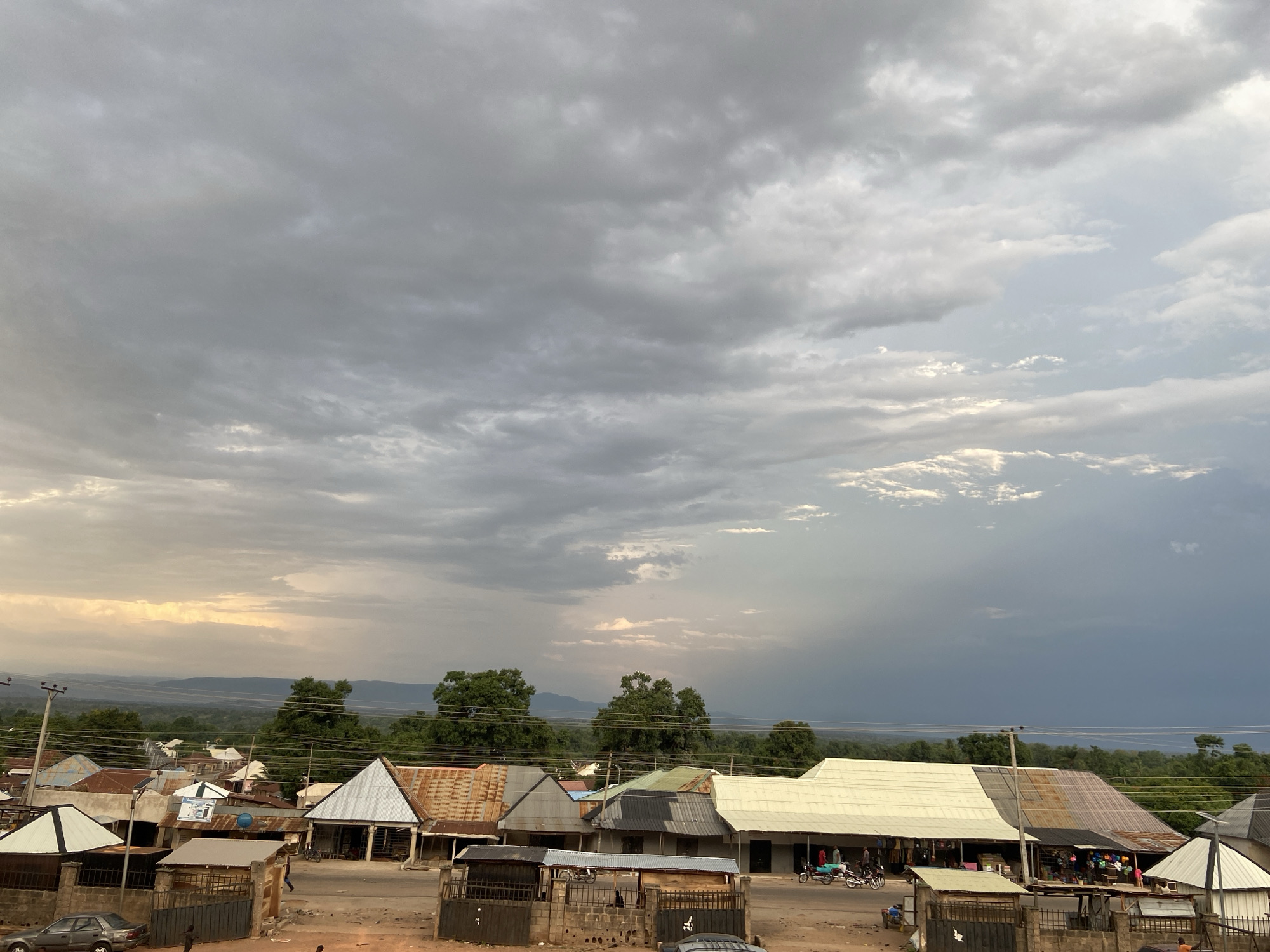 The image depicts a street scene in Tudun Wada Bibinu, Nigeria under a dramatic sky. The sky is filled with heavy, dark gray clouds, suggesting an approaching storm, with a hint of lighter clouds and sun peeking through on the right side. In the distance, rolling hills are visible.
In the foreground, there are several buildings with corrugated metal roofs, varying in color from brown to gray to white. The buildings appear to be mostly one-story structures, with some having signs and possibly serving as shops or businesses. A few people and motorcycles can be seen on the street, suggesting a somewhat active, but not bustling, atmosphere. Power lines are stretched across the scene. The street is unpaved and appears dusty. There is a car parked on the left side of the image, and brick fences and gates are located in the foreground.