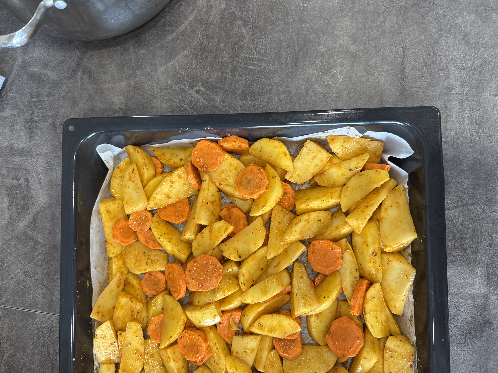 A high-angle shot showcases a black baking tray filled with seasoned potato wedges and carrot slices, prepared on a white parchment paper. The tray is positioned on a grey, textured surface. The top left corner of the image shows part of a metal pot.