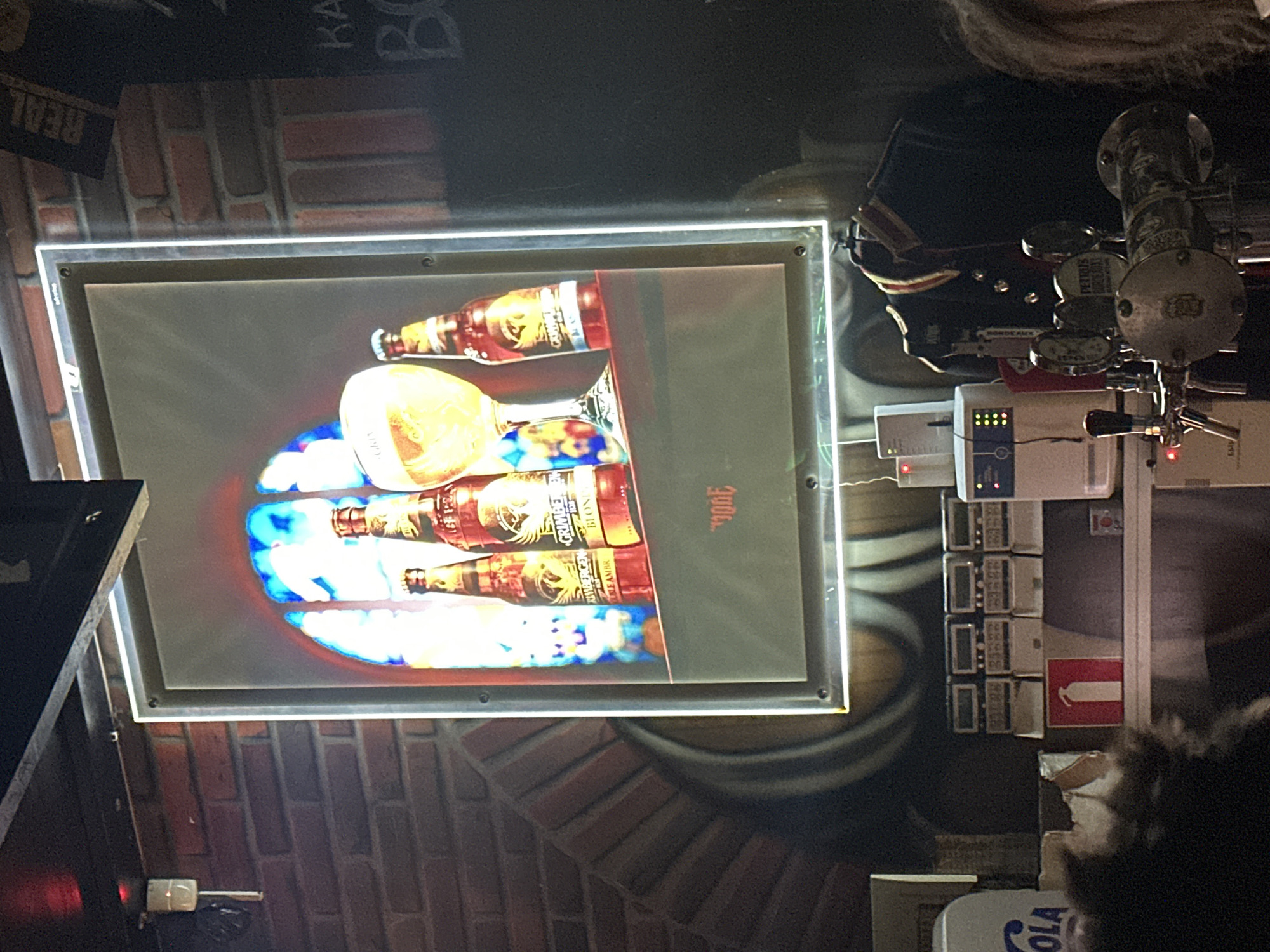The image depicts the interior of a bar, likely in Minsk, Belarus. The focal point is a large, illuminated framed advertisement featuring four bottles of Grimbergen beer and a glass of beer against a stained-glass-like background. The advertisement is hung on a brick wall. In the foreground, a bar counter is visible with several beer taps, a fire extinguisher sign, and what appear to be price displays. A dark jacket with ornate details is hanging on the wall behind the taps. The lighting is dim, with the advertisement providing a significant light source.