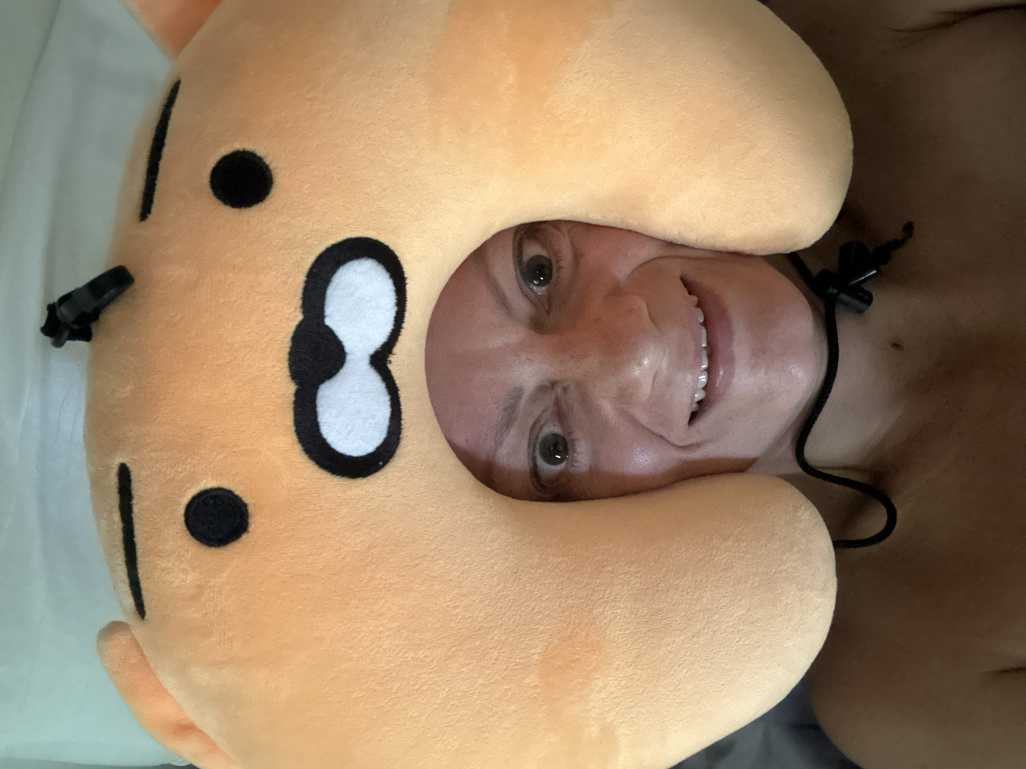 A person with light skin and short hair is seen smiling and looking directly at the camera through the head opening of an orange, cartoon-faced neck pillow. The pillow has black dots, lines, and white eye patches. A black drawstring and clasp are visible around the person's neck and upper chest. A white sheet is visible on the left side. The location is not discernable.