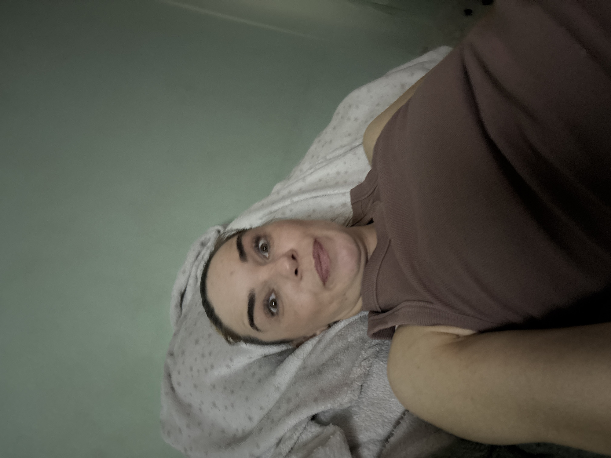 A woman with light skin, brown eyes, and brown hair pulled back is lying in bed, looking at the camera with wide eyes and a puckered mouth. She is wearing a brown tank top. A patterned blanket is behind her head, and a larger, light-colored blanket is covering her. The wall behind her is a pale green.