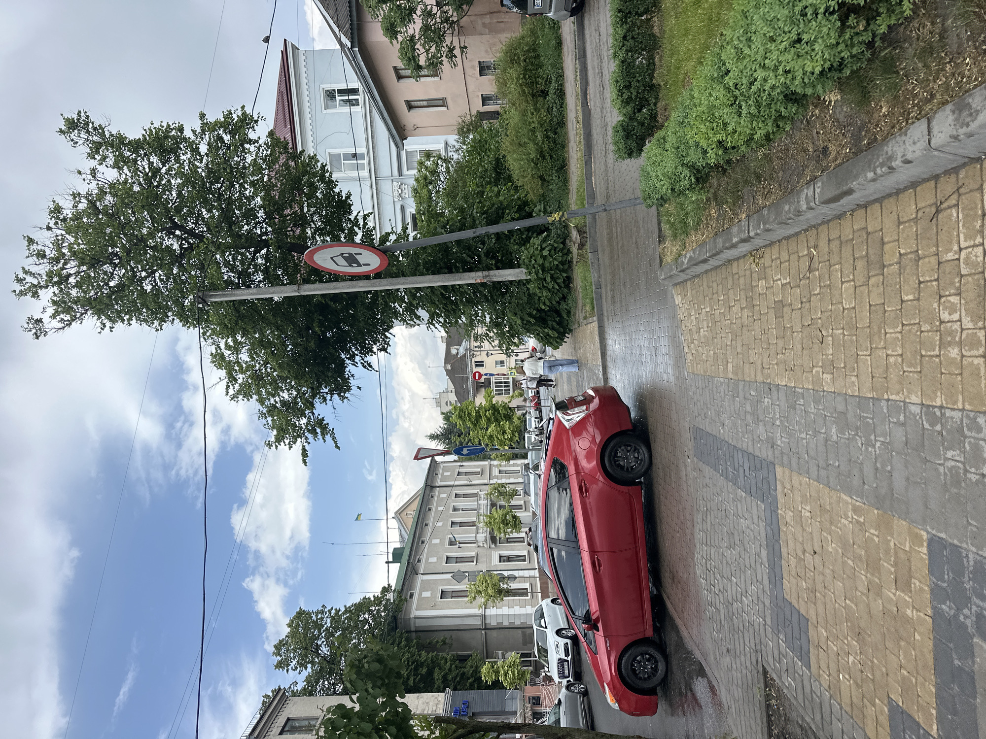 The image shows a street scene in Ternopil, Ukraine. A red car is parked on the right side of the street on a patterned brick sidewalk. A tall tree stands next to a sign prohibiting buses. In the background, several buildings are visible, including an older building with a light-colored facade. The sky is partly cloudy, with visible power lines. The weather appears to be overcast or shortly after rainfall, as the road and sidewalk are wet. Other parked cars are visible further down the street. There are a couple of persons in the middleground on the sidewalk across the street.