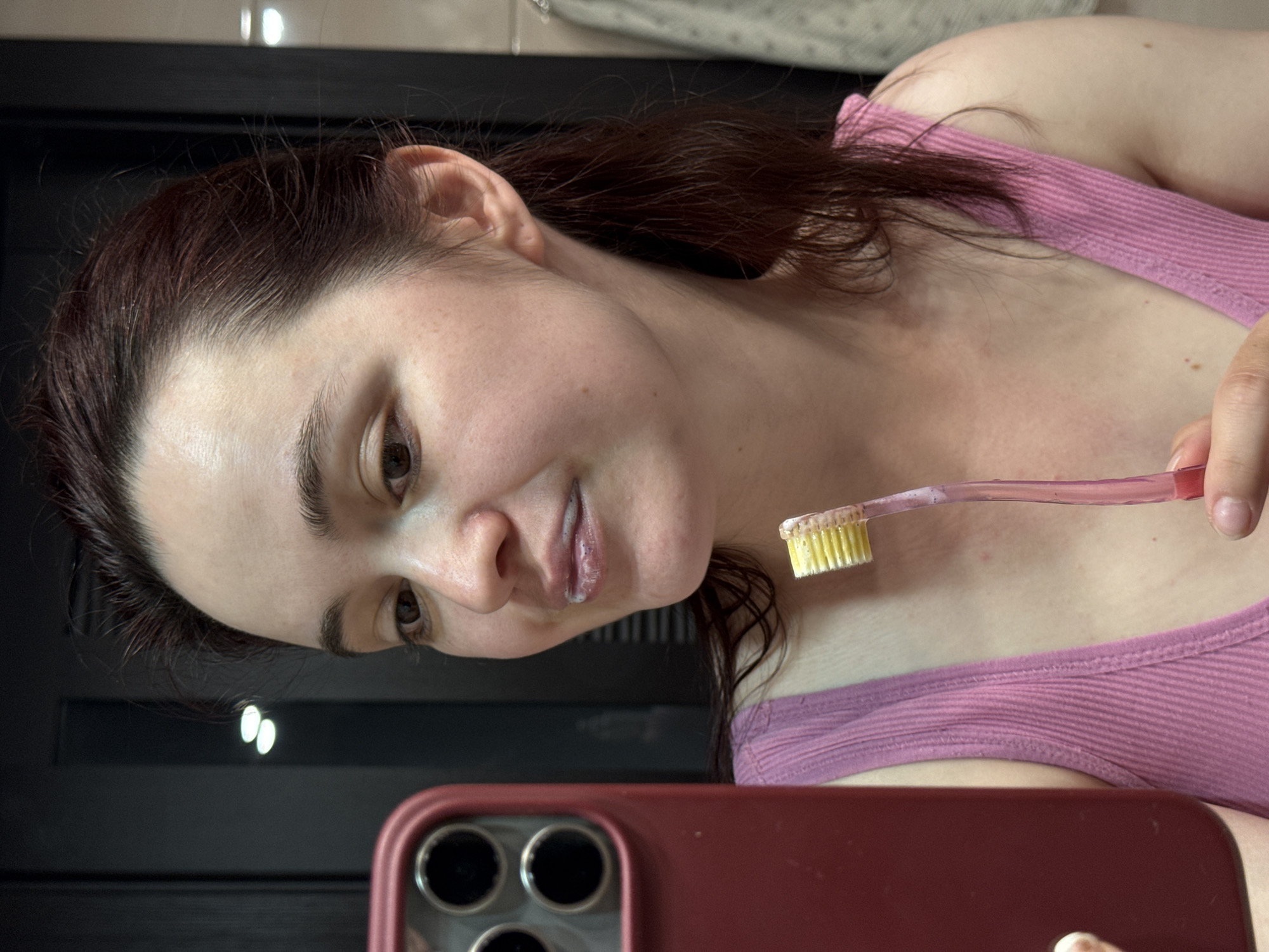 A woman is taking a selfie in what appears to be a bathroom. She is holding a smartphone with a red case in front of her. The woman is wearing a pink ribbed tank top. She is holding a toothbrush with toothpaste on it. Her hair is dark and pulled back. The background includes a dark-colored cabinet or wall and bathroom lighting. There is no information available in the image to determine if this image was taken in the city of Sochi, Russia.