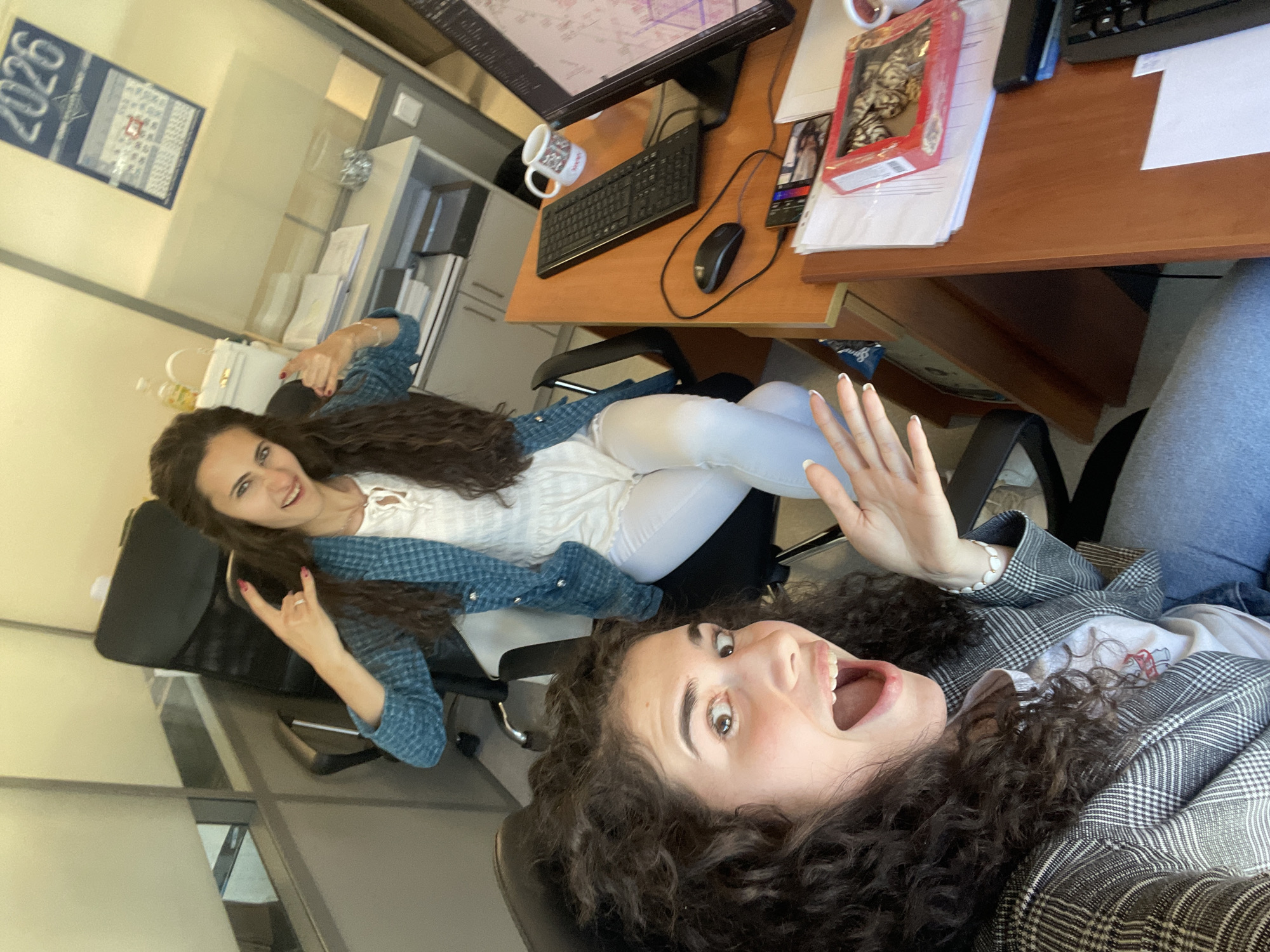 Two women are indoors, posing for a selfie. The foreground woman is sitting in a black office chair, looking directly at the camera with an open mouth expression, raising her hand. She wears a plaid blazer over a white T-shirt. The background woman is sitting in another black office chair, throwing up a "rock on" sign, wearing a white shirt, white jeans, and a denim jacket. The background reveals an office setting with a desk, computer, calendar (with “2026” visible), and a box of chocolates. The location is Sofia, Bulgaria.