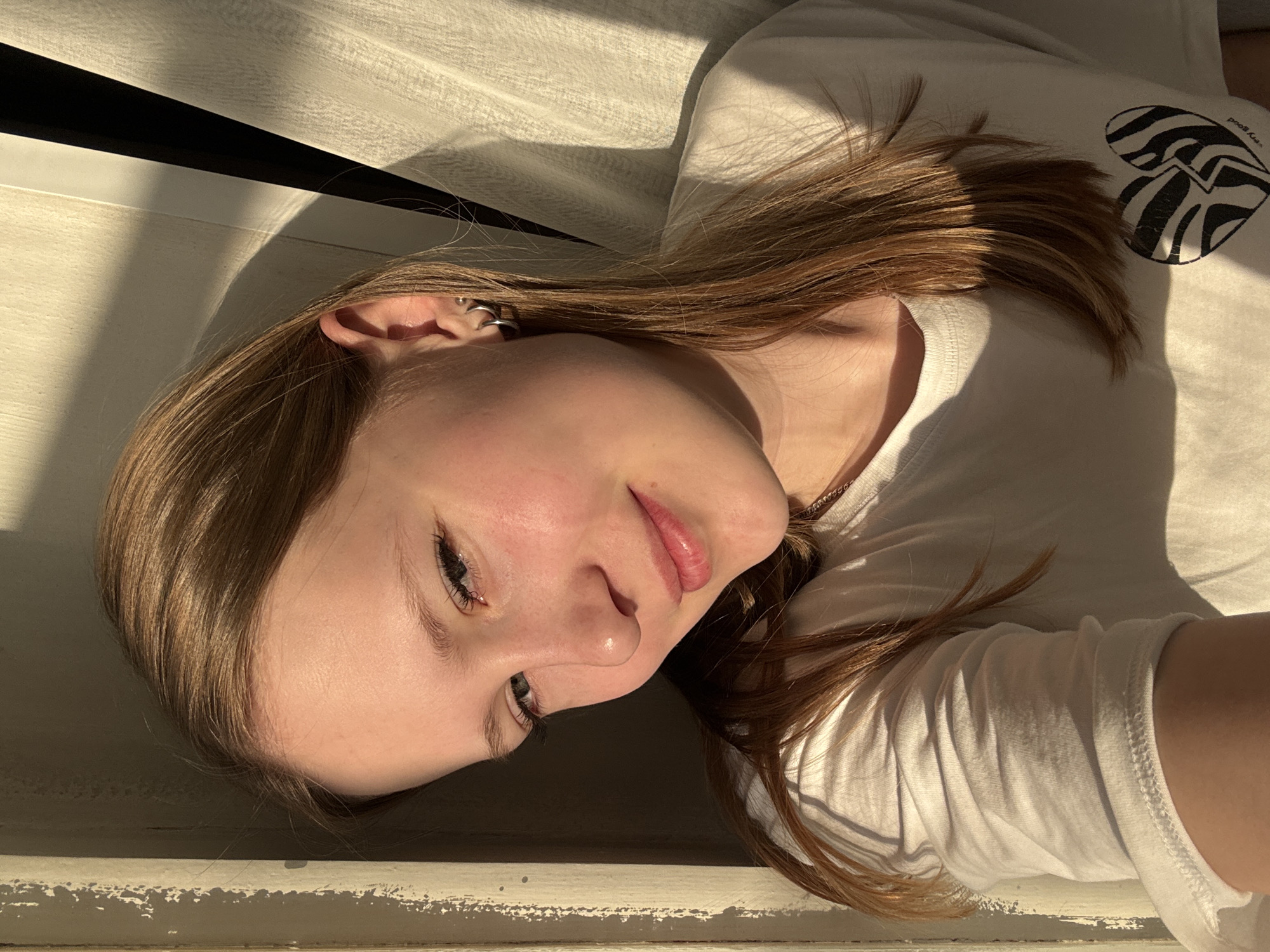 A young female with long, reddish-brown hair is depicted in a close-up, rotated portrait, seemingly lying down next to a window indoors in Chaikovsky, Russia. She wears a white short-sleeved top with a black and white zebra-print heart graphic on the upper right side, accompanied by the inverted text "very good" above it. Her left ear features multiple small hoop earrings. Strong, warm sunlight illuminates her face and hair from the side, creating distinct shadows across the light-colored surface she is resting on and the white fabric of her top. Her expression is soft and relaxed, with a slight, gentle smile. The background consists of a light-colored, possibly painted wooden window sill or ledge showing some wear, above which are prominent dark shadows and horizontal lines, likely from window blinds or a textured wall. The intense light and long shadows suggest either early morning or late afternoon sun.