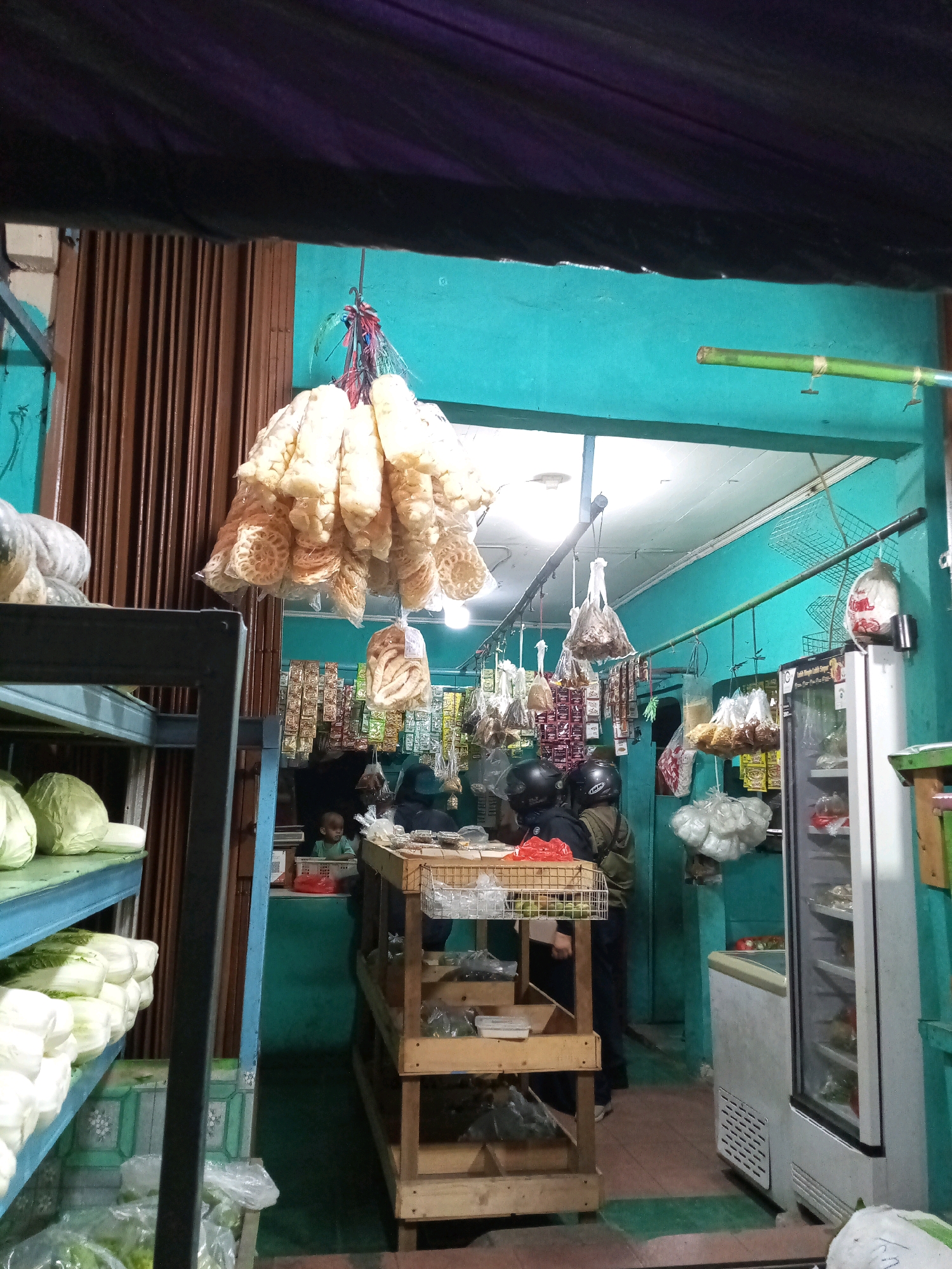 The image depicts the interior of a small store or market stall, likely in Bekasi, Indonesia, given the context. The scene is dimly lit, with turquoise walls forming the background. In the foreground, a metal shelf unit displays several cabbages and what appear to be other fresh vegetables wrapped in plastic. To the right of the shelves, a wooden display rack holds various packaged goods and some items in baskets. A white refrigerator with a glass door is positioned further back, showing some contents. Hanging from the ceiling are numerous bunches of dried snacks, possibly crackers or chips, wrapped in clear plastic. Two individuals wearing helmets are seen in the mid-ground, one facing away from the camera, and another partially obscured. Another person, a young child, is visible in the background behind a counter. The overall impression is a functional, perhaps family-run, establishment selling food items.