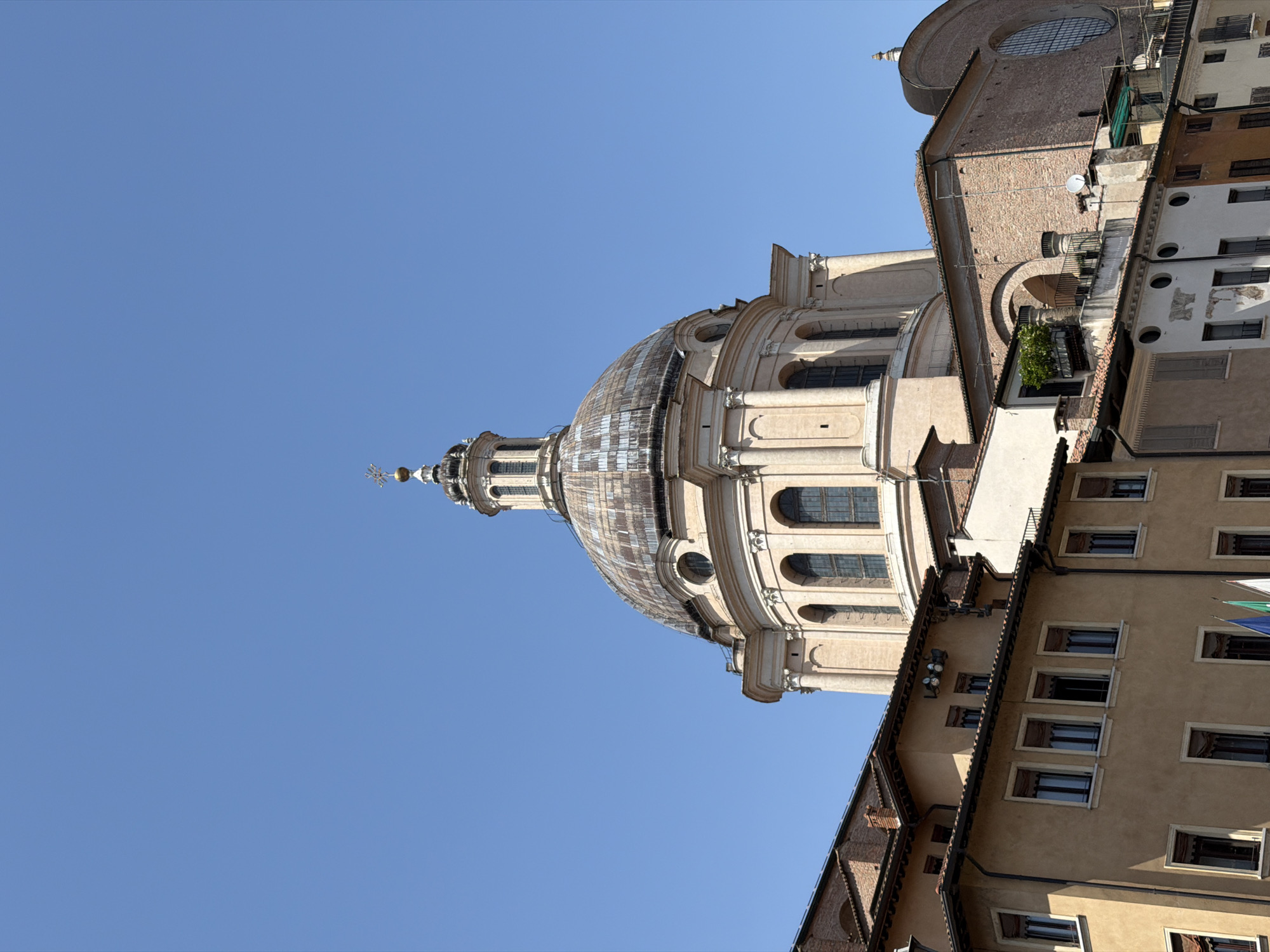 The image shows the dome of the Basilica di Sant'Andrea in Mantua, Italy, viewed from below against a clear blue sky. The dome's architectural details are visible, including its windows, columns, and ornamentation. Adjacent to the basilica, the facades of buildings with windows and other architectural elements are also seen. The image is taken from a low angle, emphasizing the height of the basilica and the surrounding buildings.