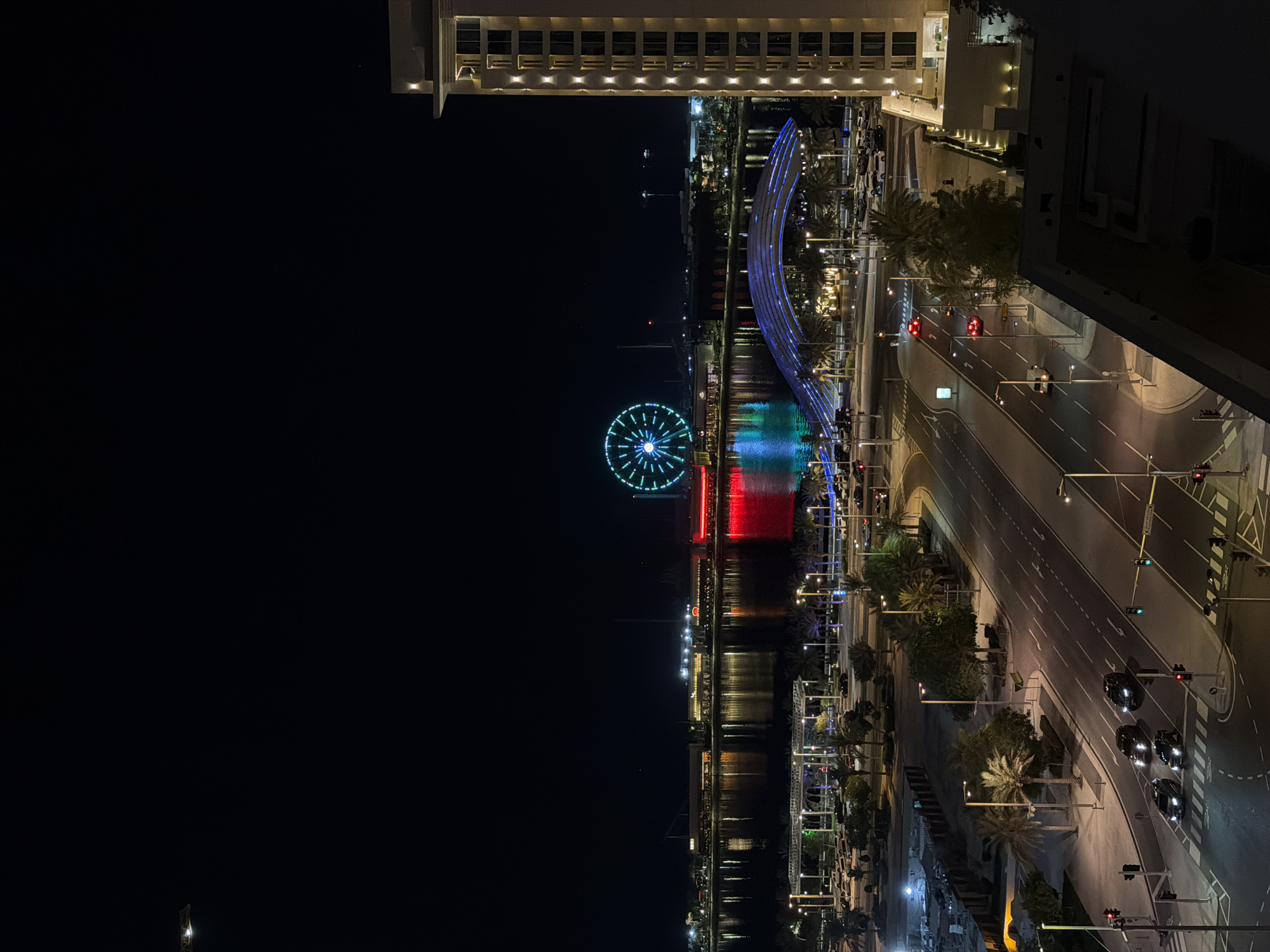 An elevated, night-time, vertical, outdoor, full shot depicts a cityscape with a Ferris wheel. A tall, modern, tan-colored building occupies the right side of the frame. Its facade is punctuated by regularly spaced, illuminated vertical lines. The building's roof is visible in the lower right corner of the frame. In the background, a large, brightly lit Ferris wheel displays a green and blue illuminated pattern. It is situated across a body of water that reflects the lights from buildings and structures along its edge, including a red illuminated waterfront promenade. A blue, arched bridge spans the water in front of the Ferris wheel. Below the bridge, palm trees and streetlights line a wide, multi-lane road where several cars are driving, their headlights and taillights illuminated. Traffic lights are visible at intersections. In the middle ground, a parking lot with numerous vehicles is illuminated by overhead lights and palm trees. A dark, unlit area is visible in the lower left corner.