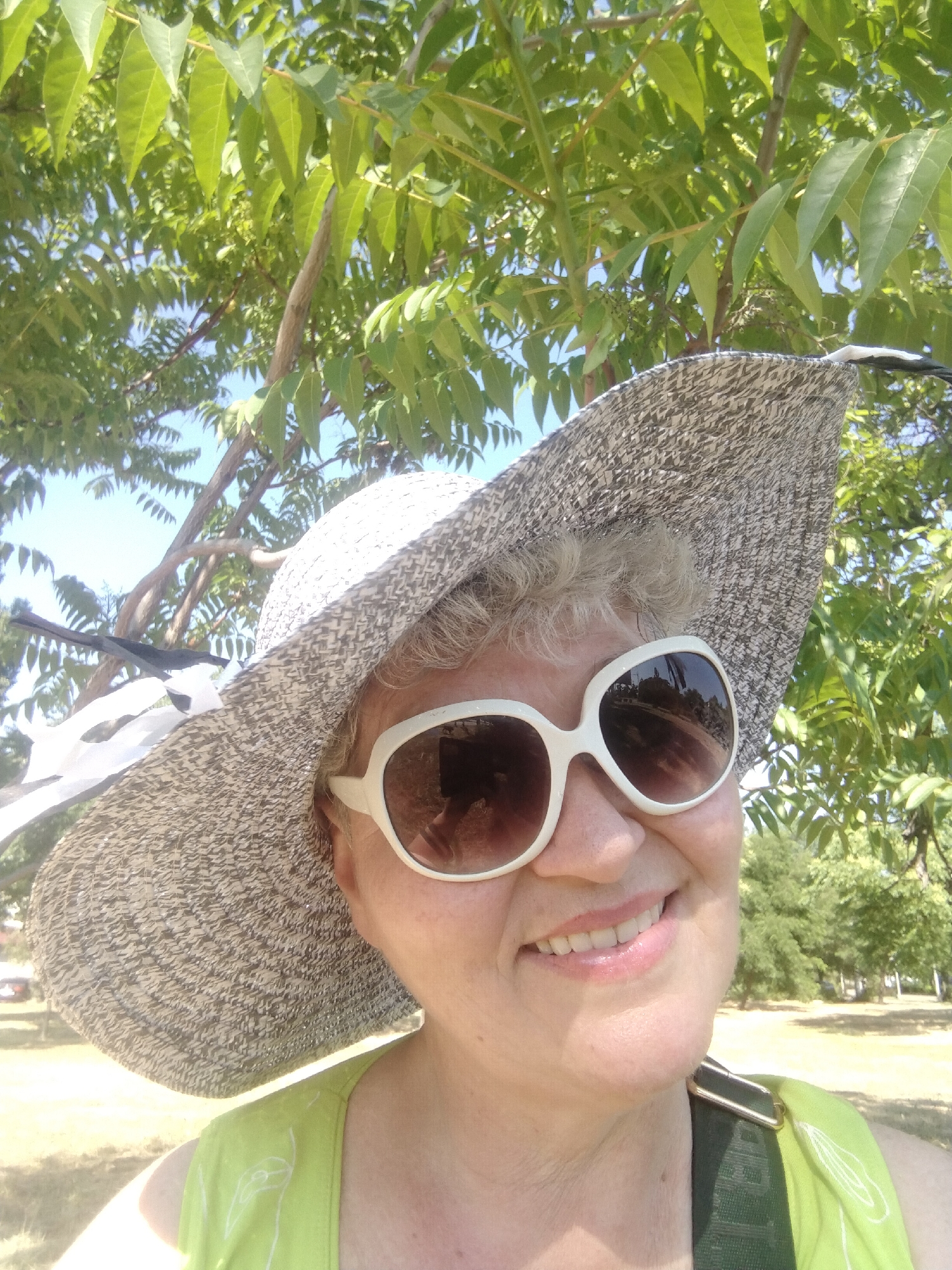 A woman is taking a selfie outdoors, likely in a park, on a sunny day. She is wearing a wide-brimmed hat and white-rimmed sunglasses, which reflect the surroundings. The background consists of green foliage from a tree canopy and the sky, suggesting an outdoor setting like a park or garden. Her expression is a slight smile. The image is framed from the chest up. The location in the city of Horishni Plavni, Ukraine is not discernible from the provided image.