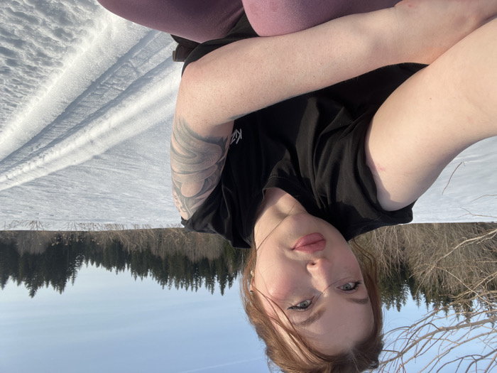 An adult female is depicted in a close-up, self-portrait photograph taken in a snowy, open landscape, likely in the vicinity of Pavlovka, Russia. The subject, with fair skin, reddish-brown hair pulled up, and light-colored eyes, faces the camera directly. She wears prominent eyeliner and light pink lipstick. Her attire consists of a black, sleeveless top and pink or mauve-colored pants. A large, dark-inked tattoo, featuring a swirling or serpentine design, is visible on her left upper arm. The background reveals an expanse of white snow under a clear, pale blue sky. Parallel tracks, consistent with skis or a sled, are imprinted in the snow to the right of the subject. Bare deciduous trees and bushes populate the mid-ground, transitioning to a dense line of dark evergreen trees in the distant background.