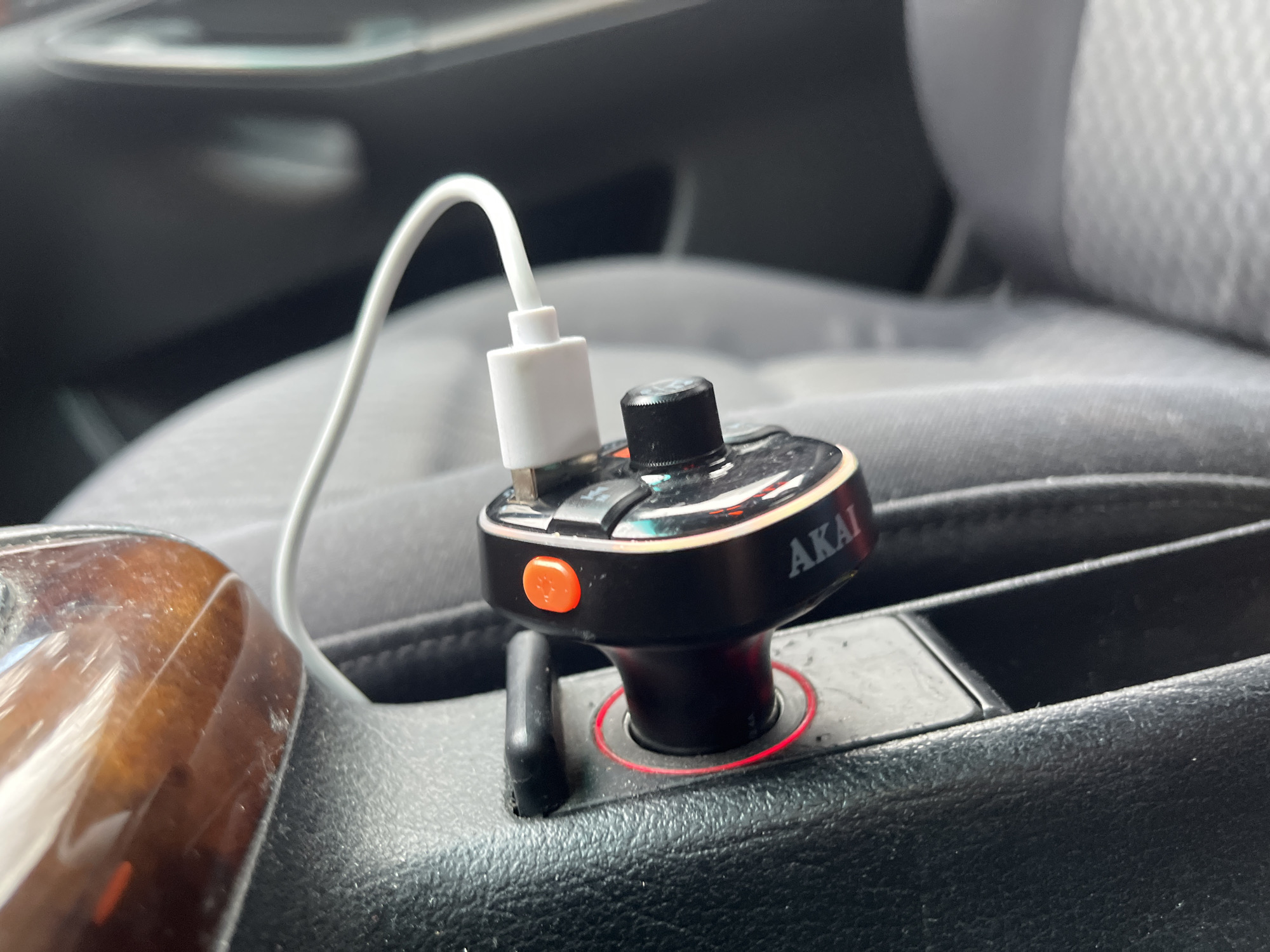 The image shows an Akai-branded FM transmitter plugged into the cigarette lighter socket of a car. A white USB cable is connected to the device. The device is located in the center console of the car, next to a wood-grain trim panel. The car's interior features dark gray upholstery. The location within the city of Sofia, Bulgaria, cannot be determined from the given image.