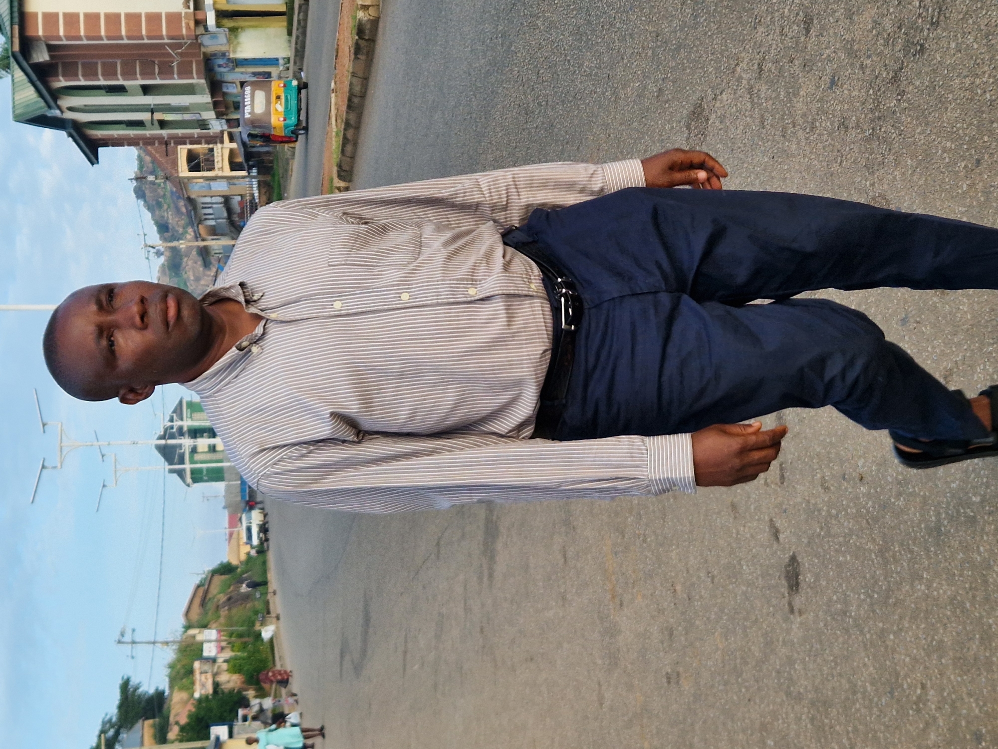 An overcast, daytime, eye-level, full-body shot shows a Black man walking towards the camera on a paved road in Kabong, Nigeria. The man, who is balding, wears a light brown striped button-down shirt, a black belt, and dark blue pants. His left arm is bent at the elbow and his right arm is slightly extended. He is wearing black sandals. The road is gray and has tire marks on it. The man is walking on the right side of the road. In the background, there are buildings and a few people walking on the sidewalk. A yellow and green tuk-tuk is driving in the street in the background. The sky is light blue and there are some clouds.