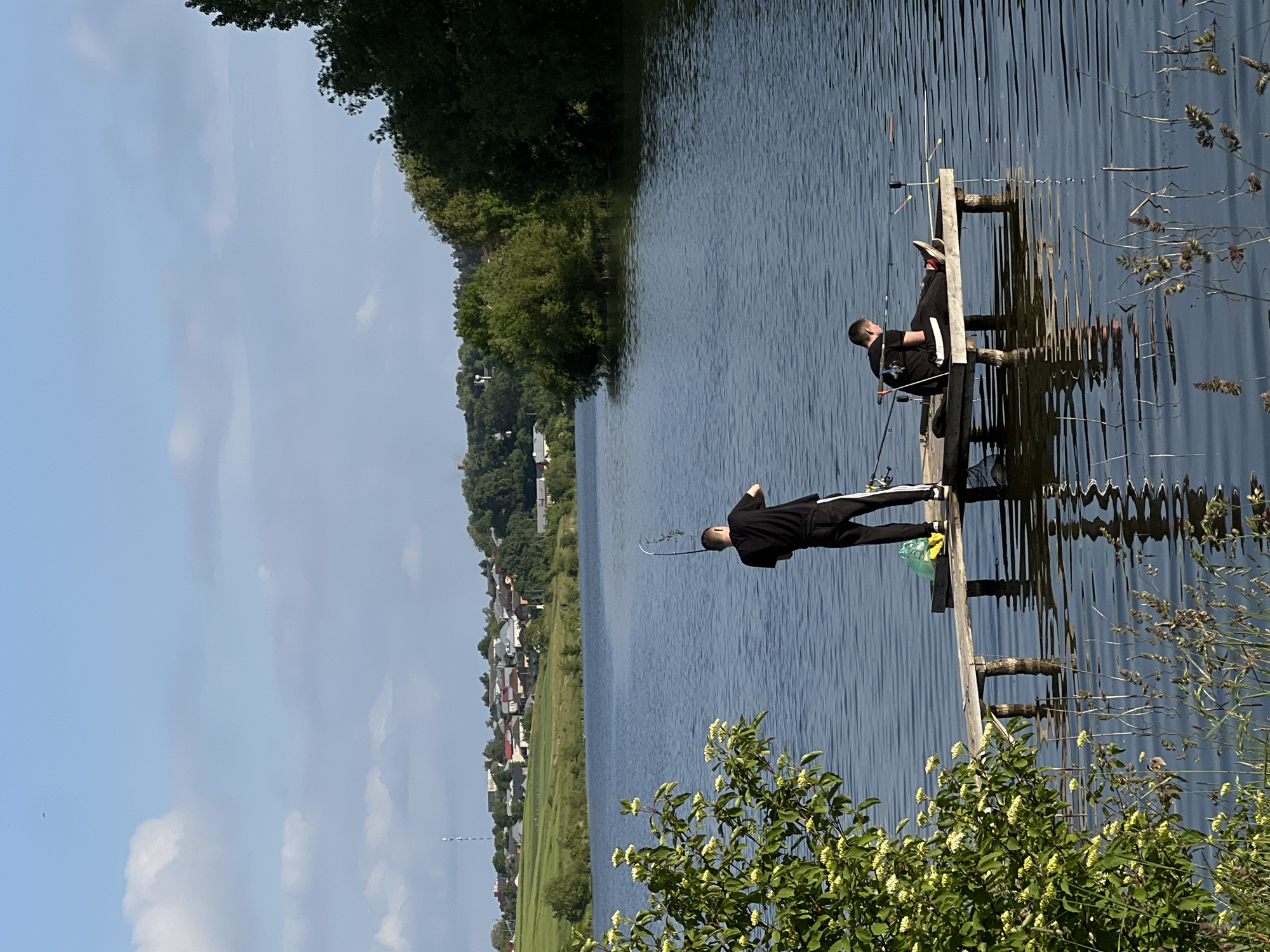 The image captures a peaceful daytime scene of two men fishing from a wooden pier extending into a calm body of water, likely a lake or wide river. The sky above is bright blue with scattered white clouds, suggesting clear and sunny weather.
In the mid-ground, two men dressed in black sportswear are positioned on the pier. One man is standing closer to the camera, actively holding a fishing rod. Further along the pier, the second man is seated, with multiple fishing rods set up around him, and a yellow plastic bag is visible nearby. Their postures indicate a relaxed recreational activity.
The foreground on the right features a lush green bush adorned with small white or pale yellow flowers. The water shows gentle ripples and reflects the light, appearing deep blue. On the distant bank, a dense line of green trees gives way to a village or small town with houses and buildings visible amidst the greenery. A slender communication tower stands prominently in the far distance, providing a landmark.
The scene conveys a tranquil atmosphere, focusing on the fishing activity in a serene natural setting. The location is Bazaryntsi, Ukraine. No visible text is present in the image.