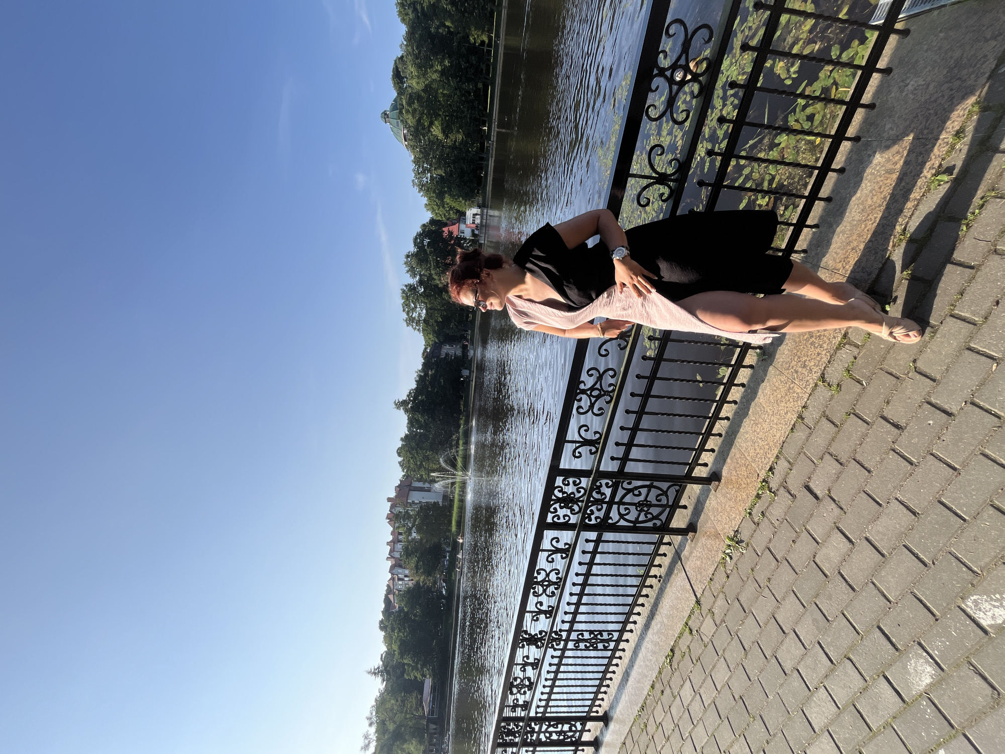A woman stands on a brick walkway next to a black metal fence overlooking a body of water. She is wearing a pink and black dress, sunglasses, and sandals. The water is calm and reflects the blue sky. In the background, there are trees, buildings, and a fountain spraying water. The scene appears to be in a park or waterfront area, possibly in Svetlogorsk, Russia.