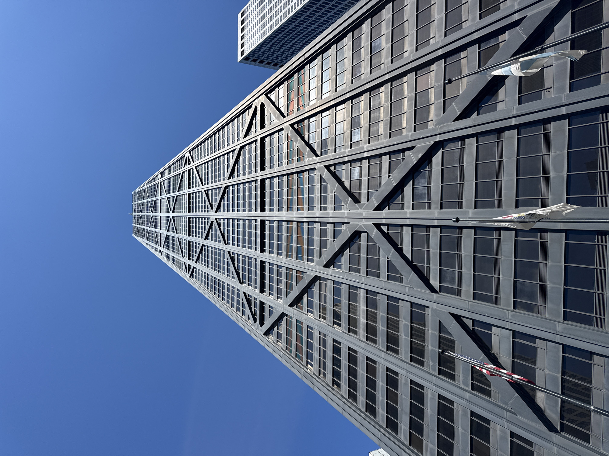This is a low-angle shot of the John Hancock Center in Chicago, USA. The building's distinctive X-braced exoskeleton dominates the frame, with its dark windows reflecting the clear blue sky. Two flags are visible, one at mid-height and another lower down, both unfurled and pointing to the right. The top of another skyscraper is visible in the upper left corner, its white facade contrasting with the dark gray of the John Hancock Center. The lighting suggests it is daytime, with the sun casting distinct shadows from the building's structural elements. There is no discernible activity taking place beyond the presence of the building itself and the flags. The image captures a clear, sunny day in Chicago, highlighting the architectural features of the iconic skyscraper.