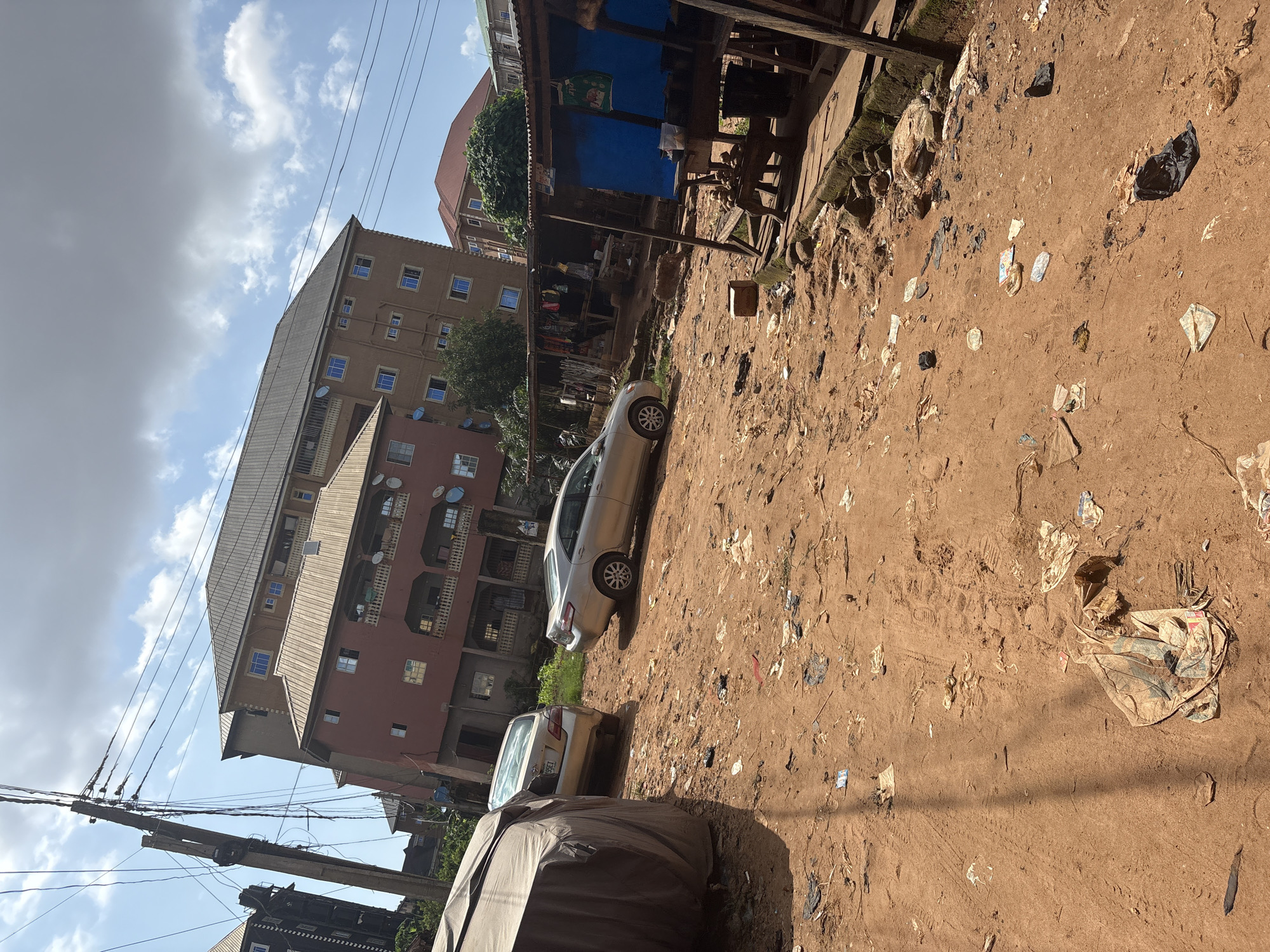 The image depicts a street scene in Nkpor, Nigeria, characterized by a dirt road littered with debris. Two silver sedan cars are parked on the right side of the road, one of which is partially covered by a grey tarp. In the background, a multi-story building with red and tan facades stands tall, adorned with satellite dishes on its balconies. To the right of the cars, a makeshift stall with a corrugated metal roof displays various items, including what appears to be food products and clothing. The sky is partly cloudy, and a utility pole with numerous wires is visible on the left side of the frame. The overall environment suggests a somewhat unkempt urban setting.